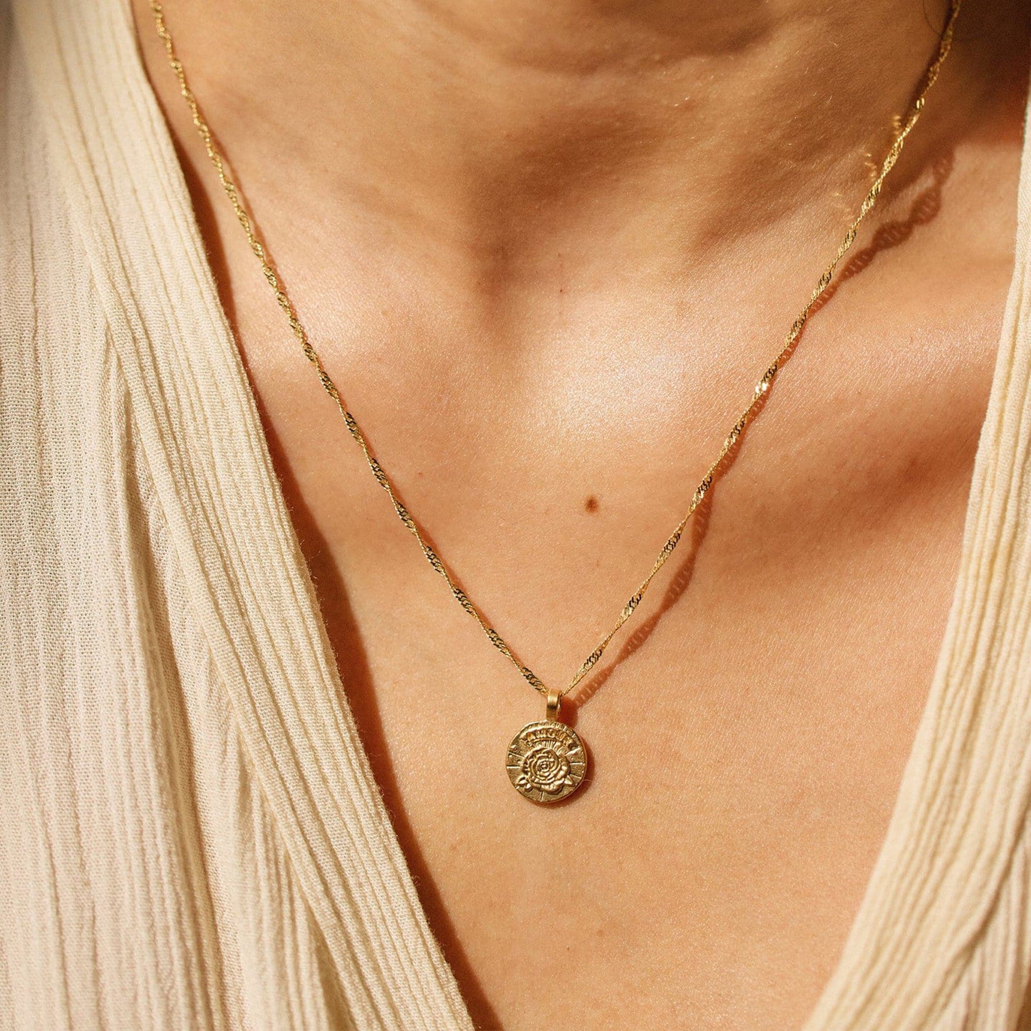 This picture is a close up of a woman’s neck. She is wearing a tan blouse. Around her neck is a gold chain with a gold circle pendent on the end. In the middle of the pendent is a gold rose. 