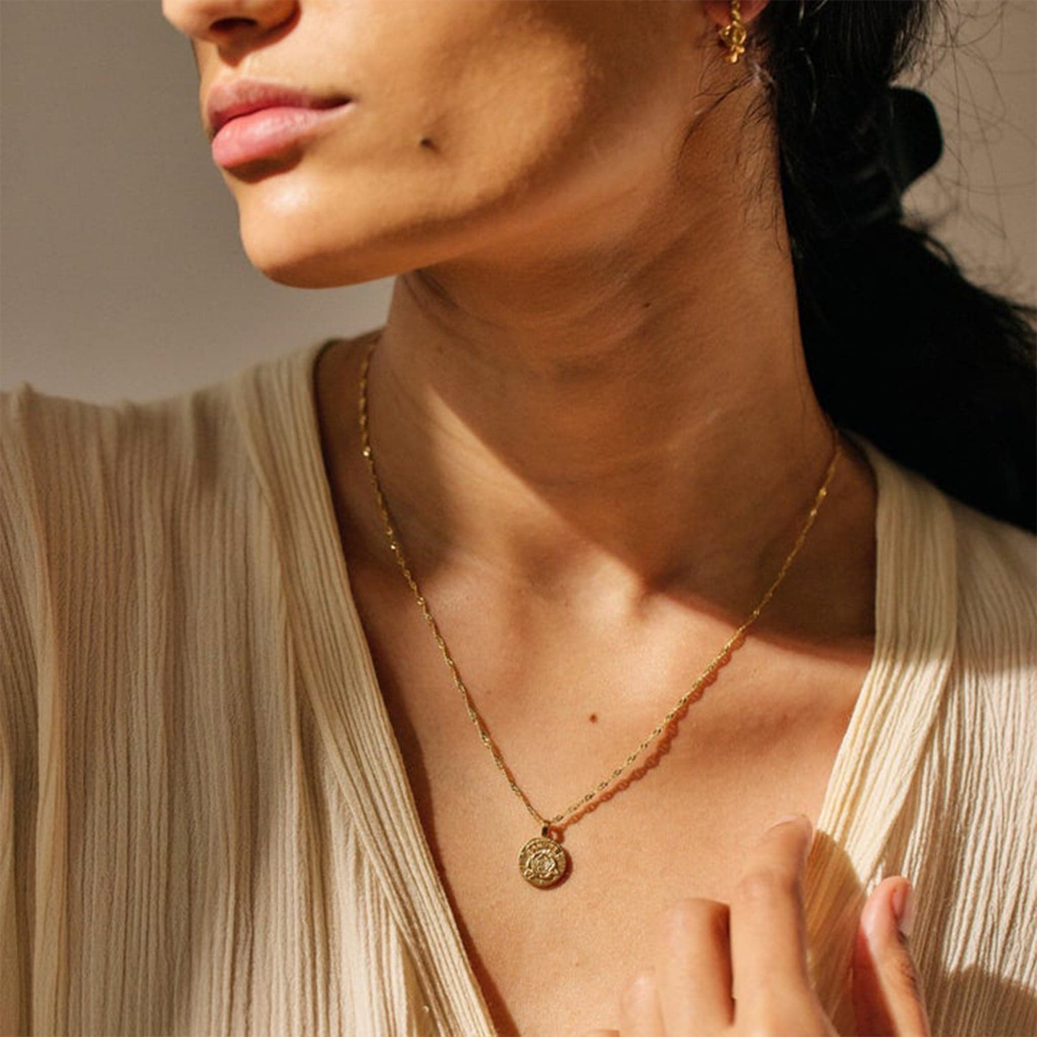 This picture is a close up of a woman’s neck. She is wearing a tan blouse. Around her neck is a gold chain with a gold circle pendent on the end. In the middle of the pendent is a gold rose. 