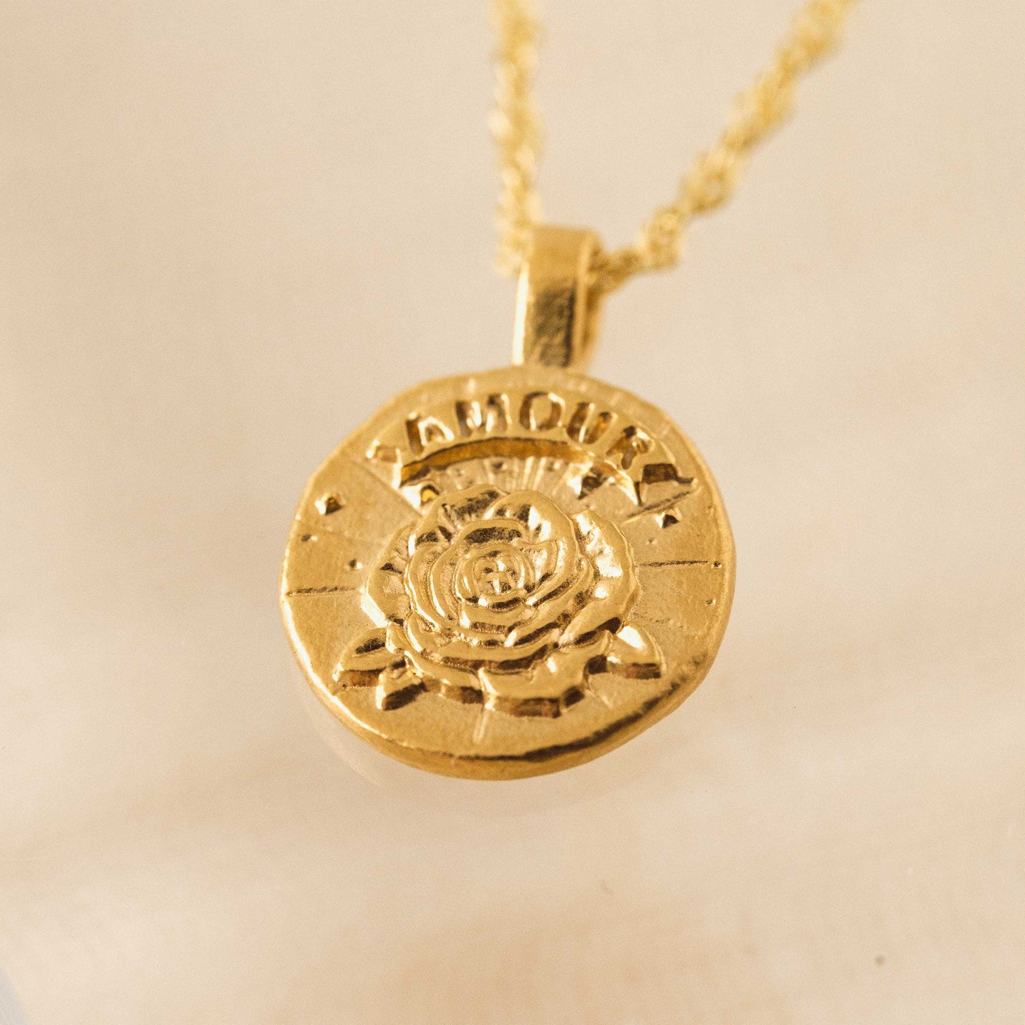 In front of a tan background is a close up of a round gold pendent on a gold chain necklace. In the middle of the pendent is a gold rose with a gold banner at the top. Etched in the banner is the word ‘Amour.&#39;