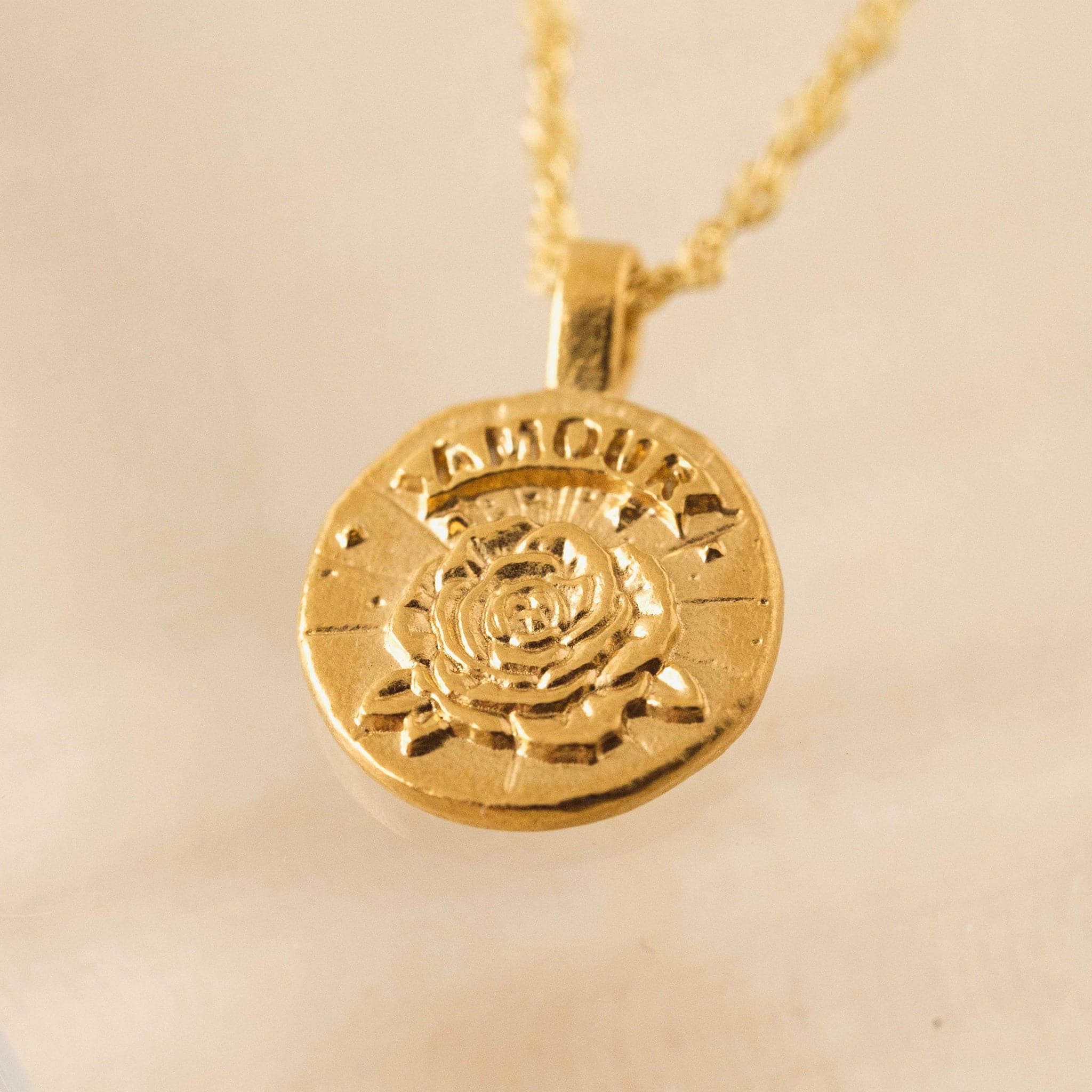 In front of a tan background is a close up of a round gold pendent on a gold chain necklace. In the middle of the pendent is a gold rose with a gold banner at the top. Etched in the banner is the word ‘Amour.'