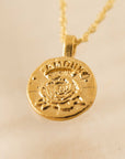 In front of a tan background is a close up of a round gold pendent on a gold chain necklace. In the middle of the pendent is a gold rose with a gold banner at the top. Etched in the banner is the word ‘Amour.'
