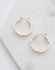 Medium sized silver hoops that have a wider frame and a thin depth. They feature a straight post and a clasp on the back top of the hoop to secure it on your ear.