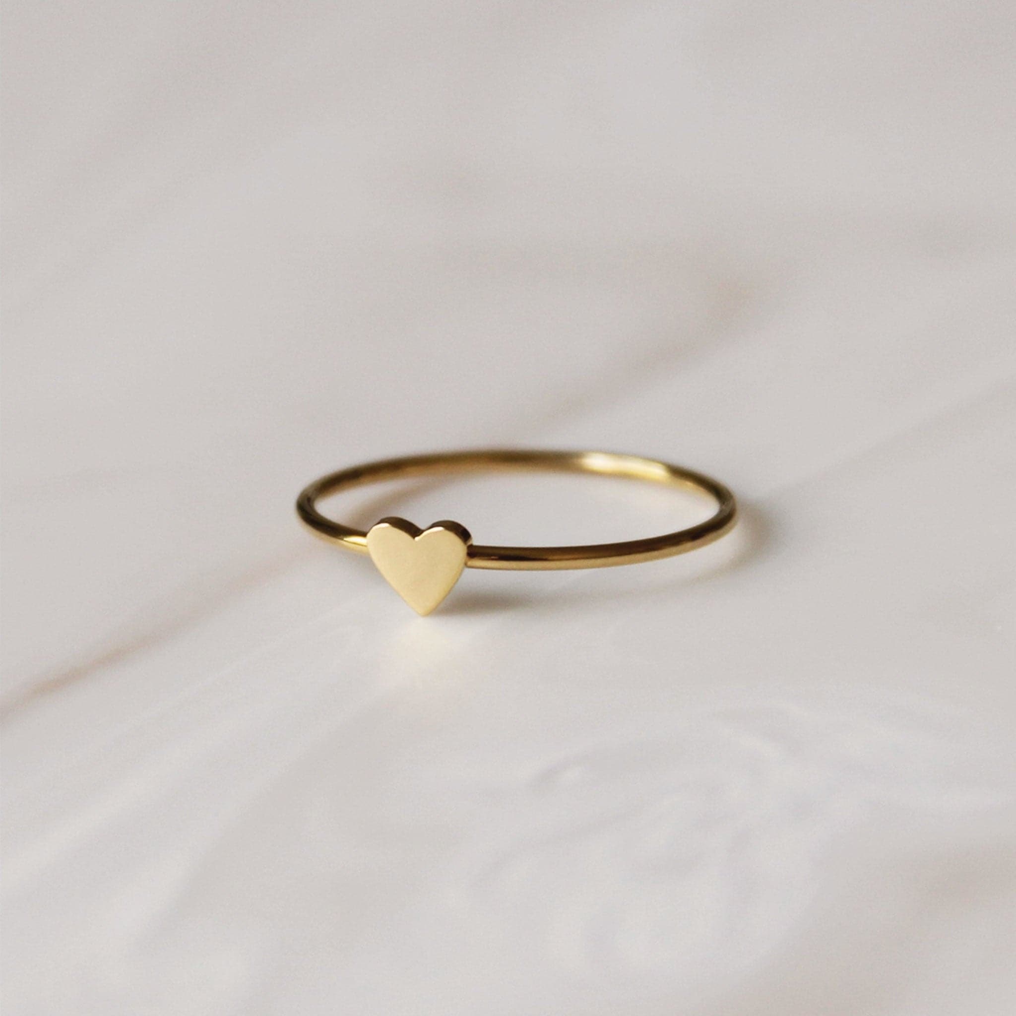 Photo of a thin gold ring with a small gold heart on it sitting on a marble surface.