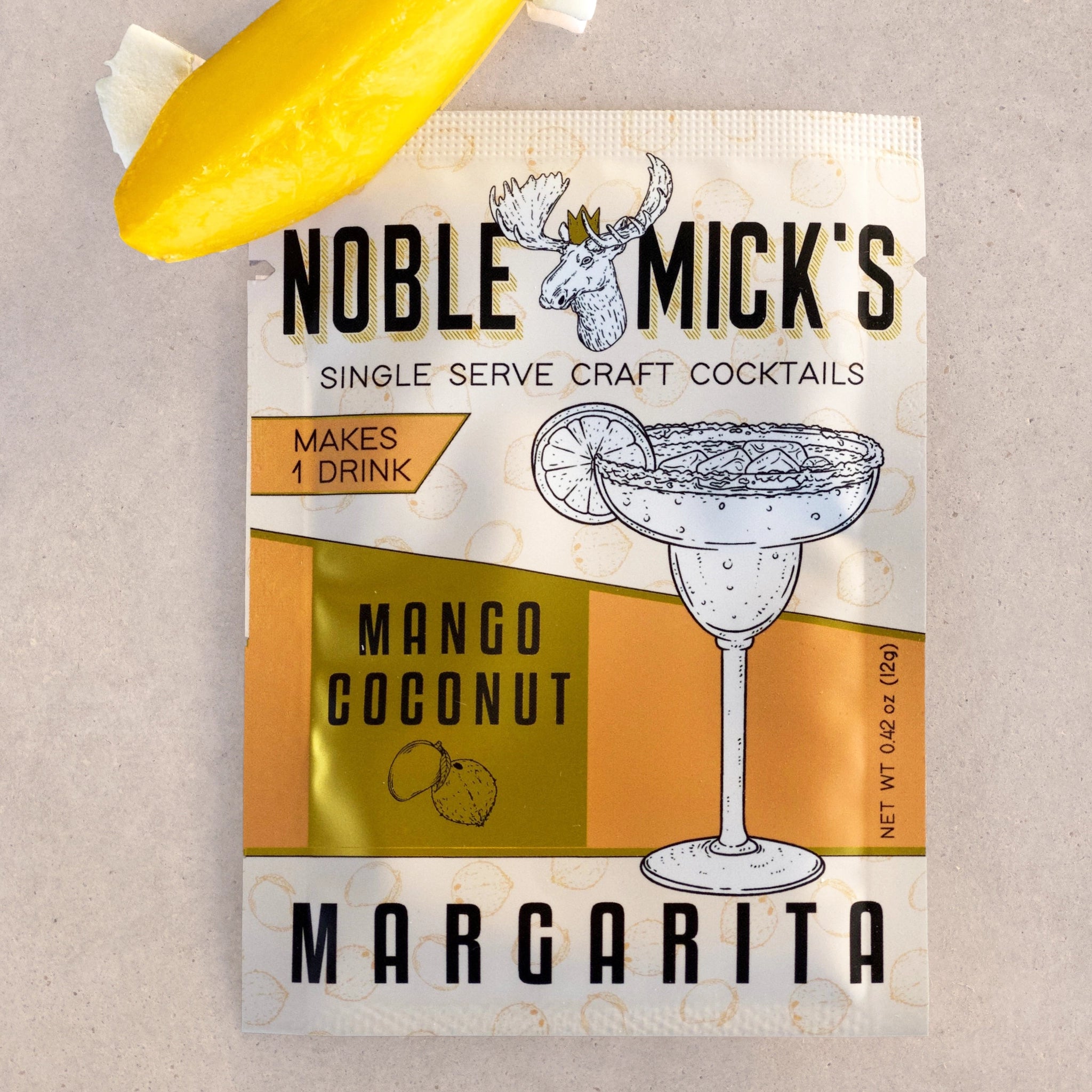 A cream and orange packet of cocktail mix that reads, &quot;Noble Mick&#39;s Single Serve Craft Cocktails Mango Coconut Margarita&quot; in black letters photographed next to a slice of mango. 