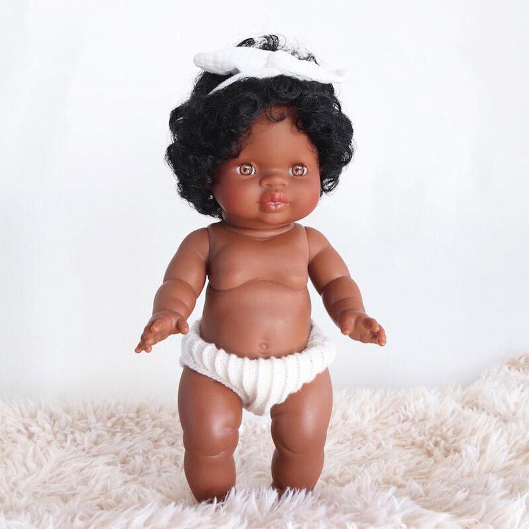 On a white background is an African American baby doll with curly hair and dressed in white knit bottoms and a white bow tie headband. 