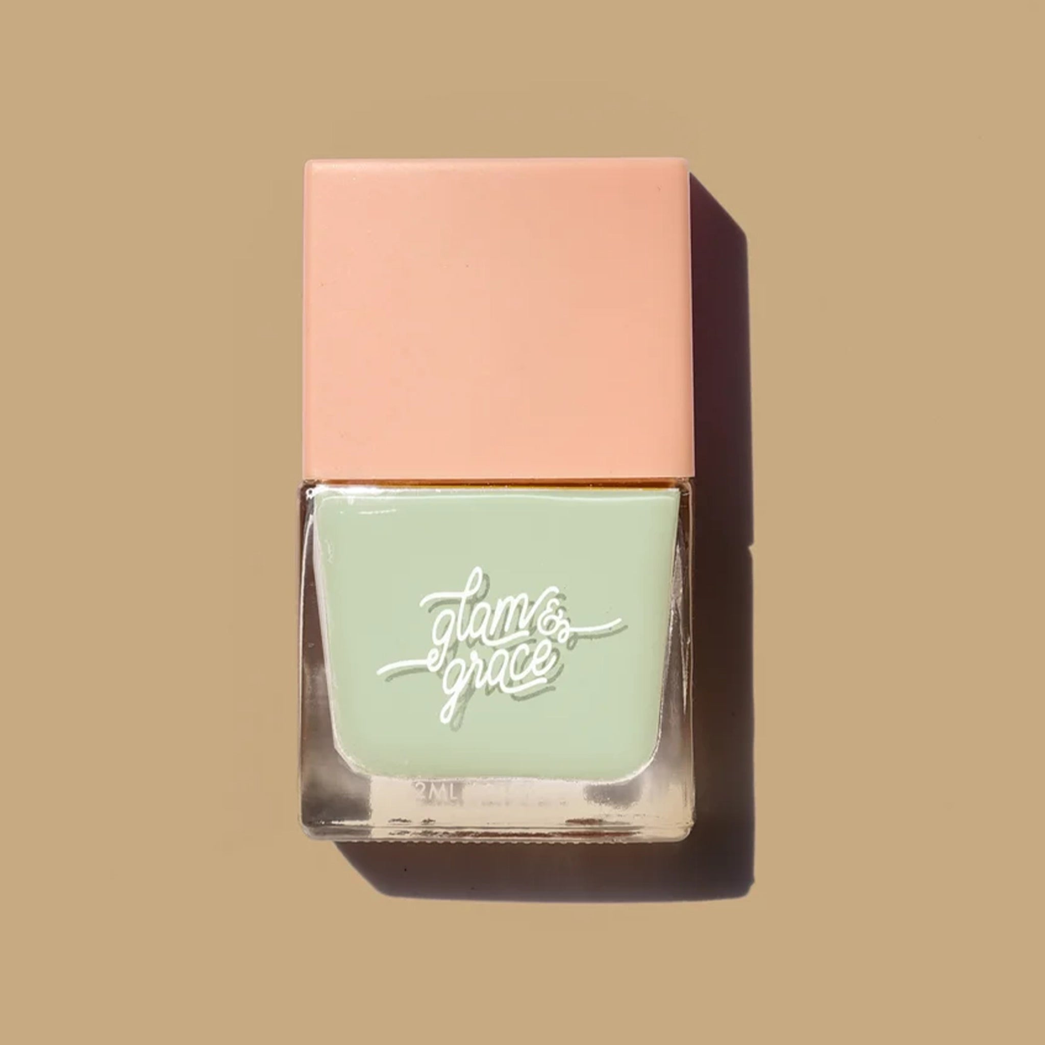 A mint green nail polish in a square glass jar with a salmon pink lid as well as white cursive text that reads, "glam &amp; grace".