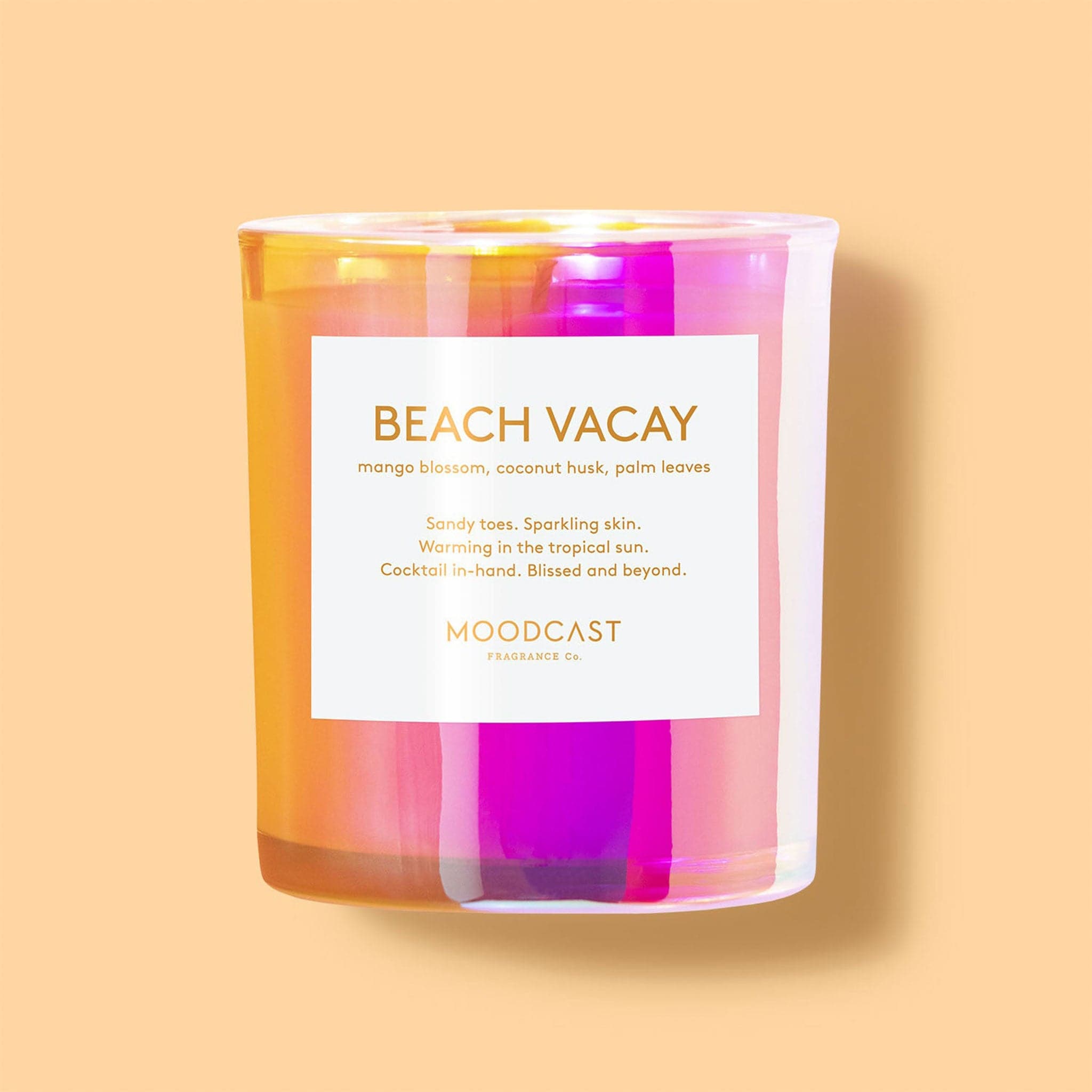 An orange and vibrant pink iridescent glass jar filled with a candle along with a white square label on the front that repeats the notes of the candle as well as the name, "Beach Vacay" in gold letters.