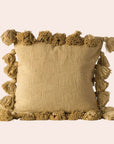 On a cream background, is a mustard yellow pillow with tassels from all alone the edges of the square pillow.