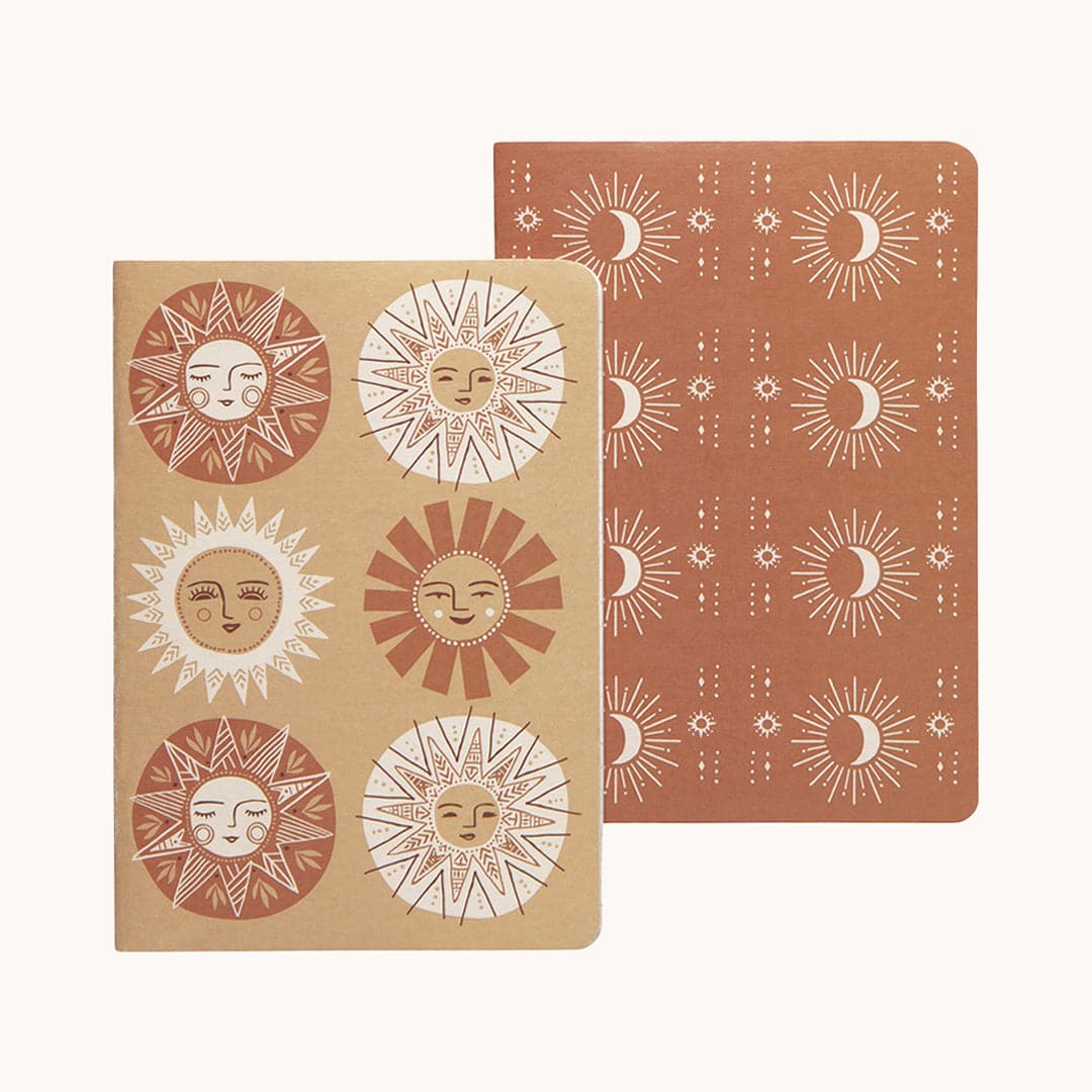 In front of a white background a tan notebook with six tan, white and rust suns on the cover. All six of the suns have a face and unique sun beams. Under is a rust notebook with a white pattern on the cover. There are crescent moons with sun beams in the pattern. 