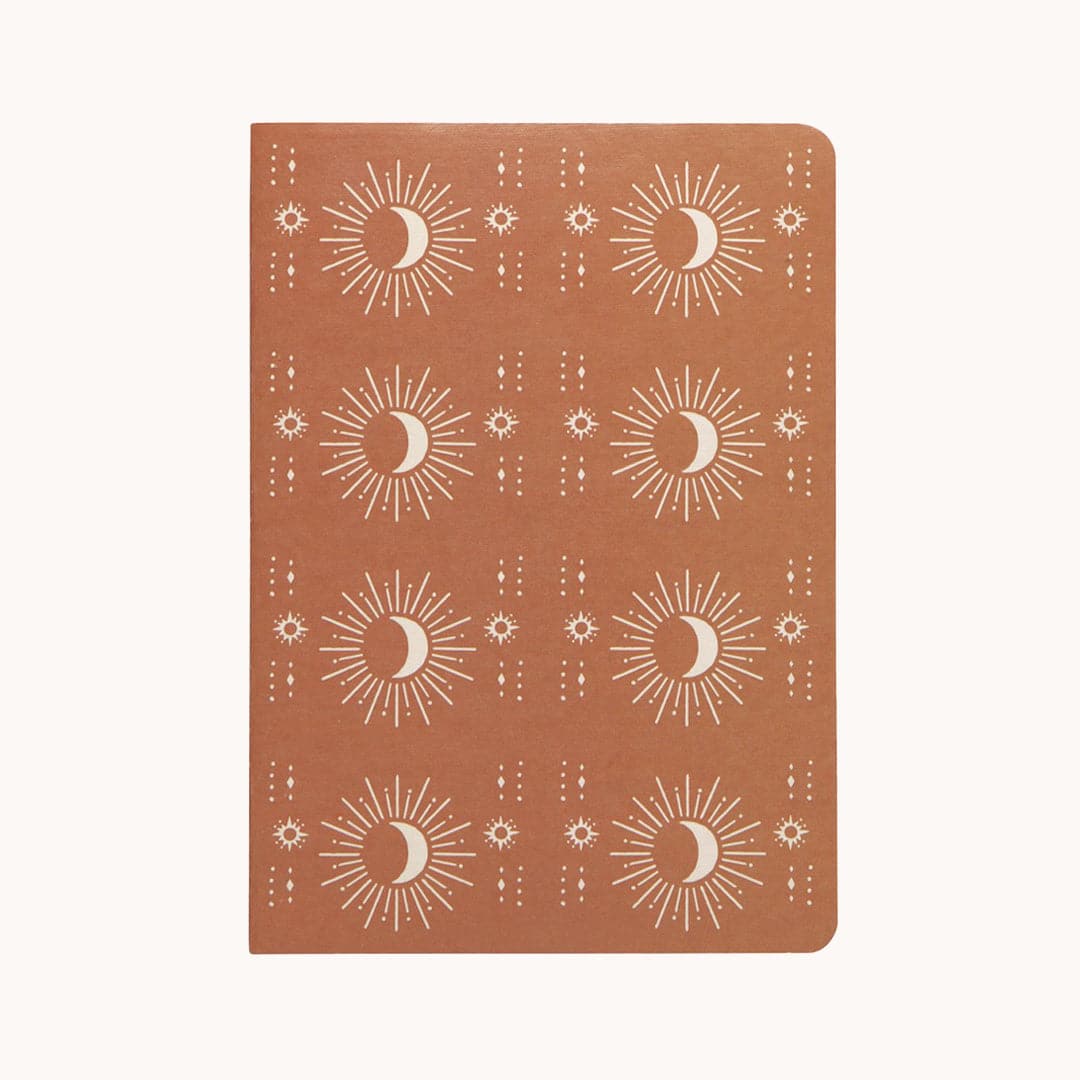 In front of a white background is a rust notebook with a white pattern on the cover. There are two vertical rows of white crescent moons with white sun beams. In between the rows are lines of white dots and circles. 