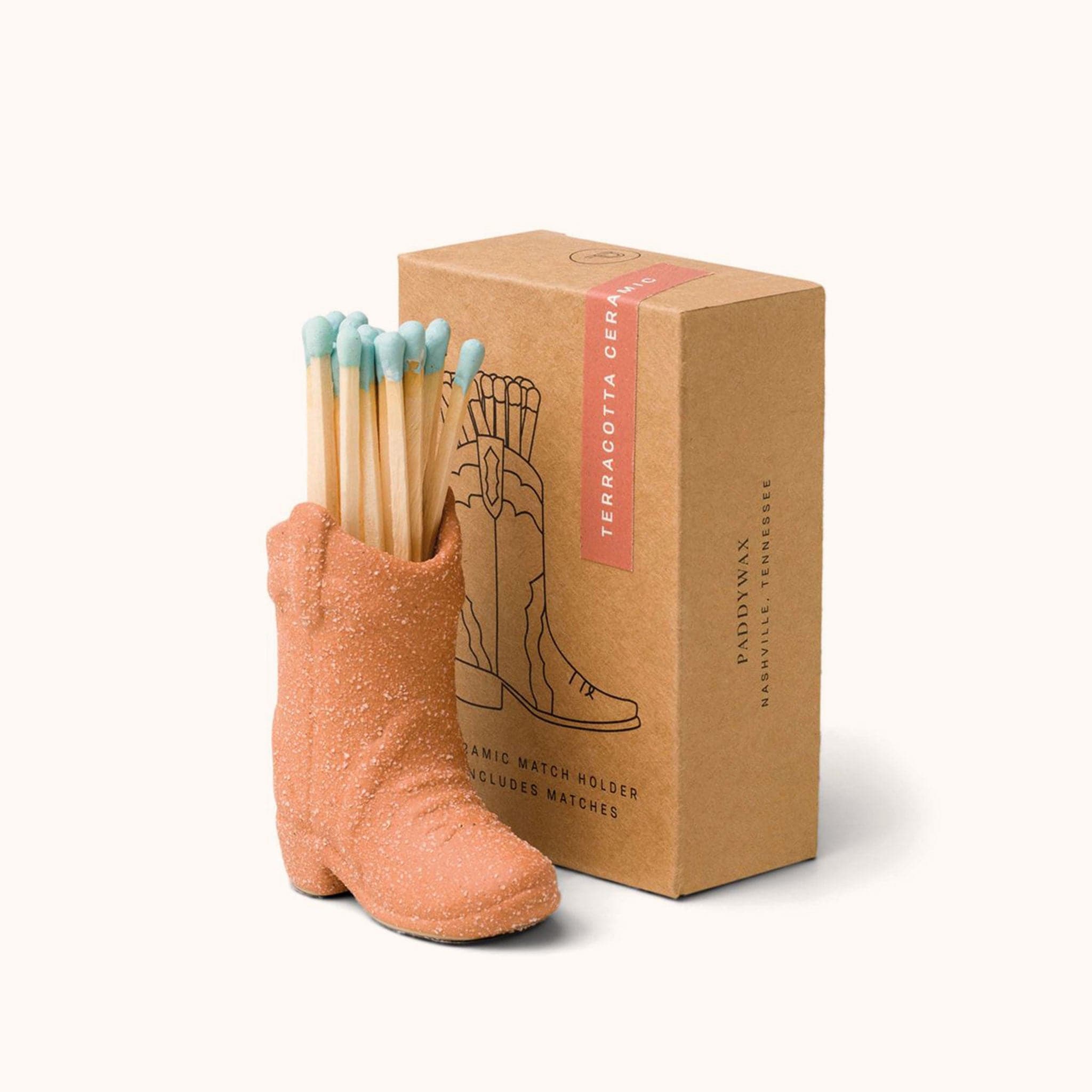 Terracotta ceramic boot match holder filled with a bundle of blue dipped matches. The holder is positioned next to kraft brown packaging.