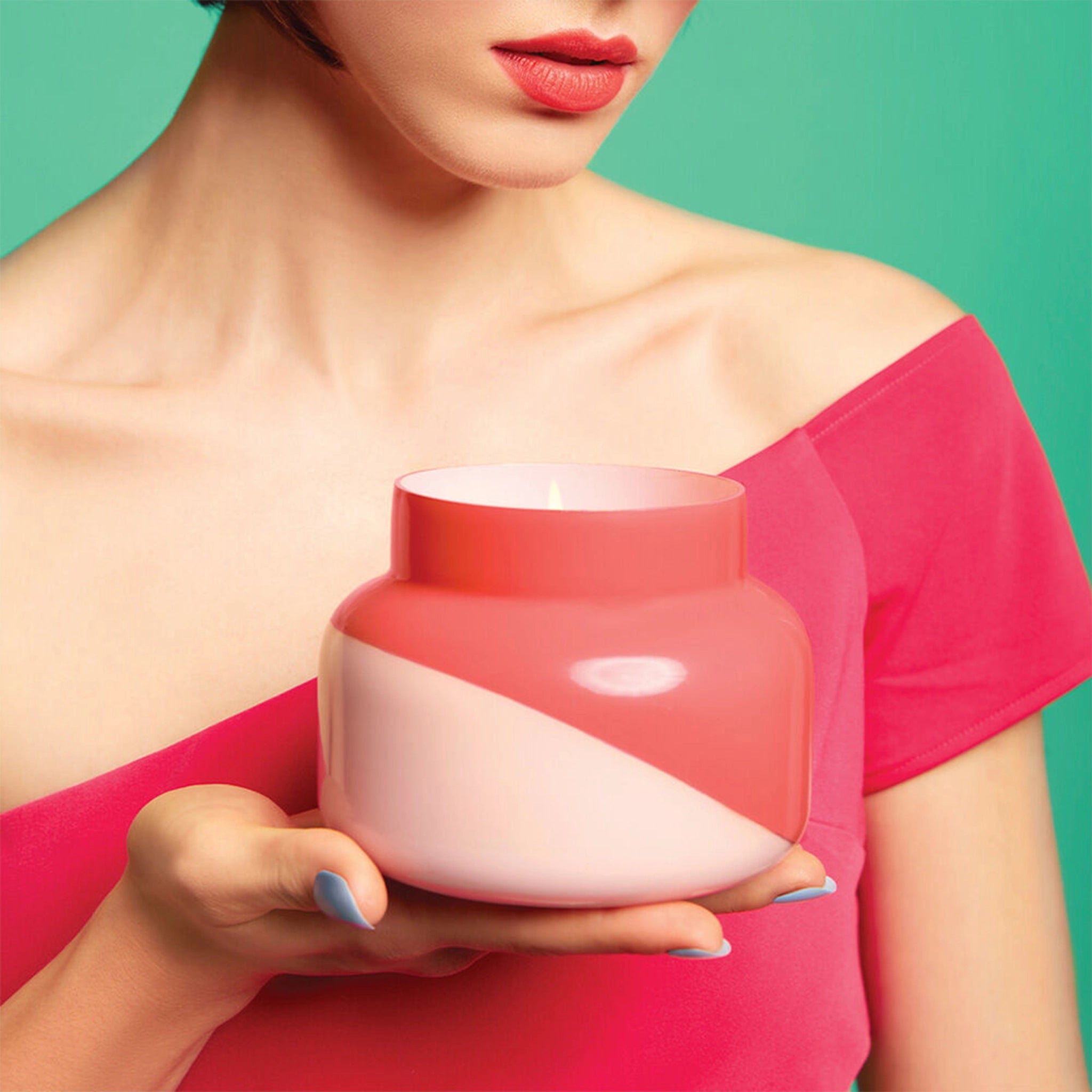 A glass candle jar with a two toned design featuring a light pink and a darker pink that is split diagonally being held by a model wearing a hot pink off the shoulder dress.