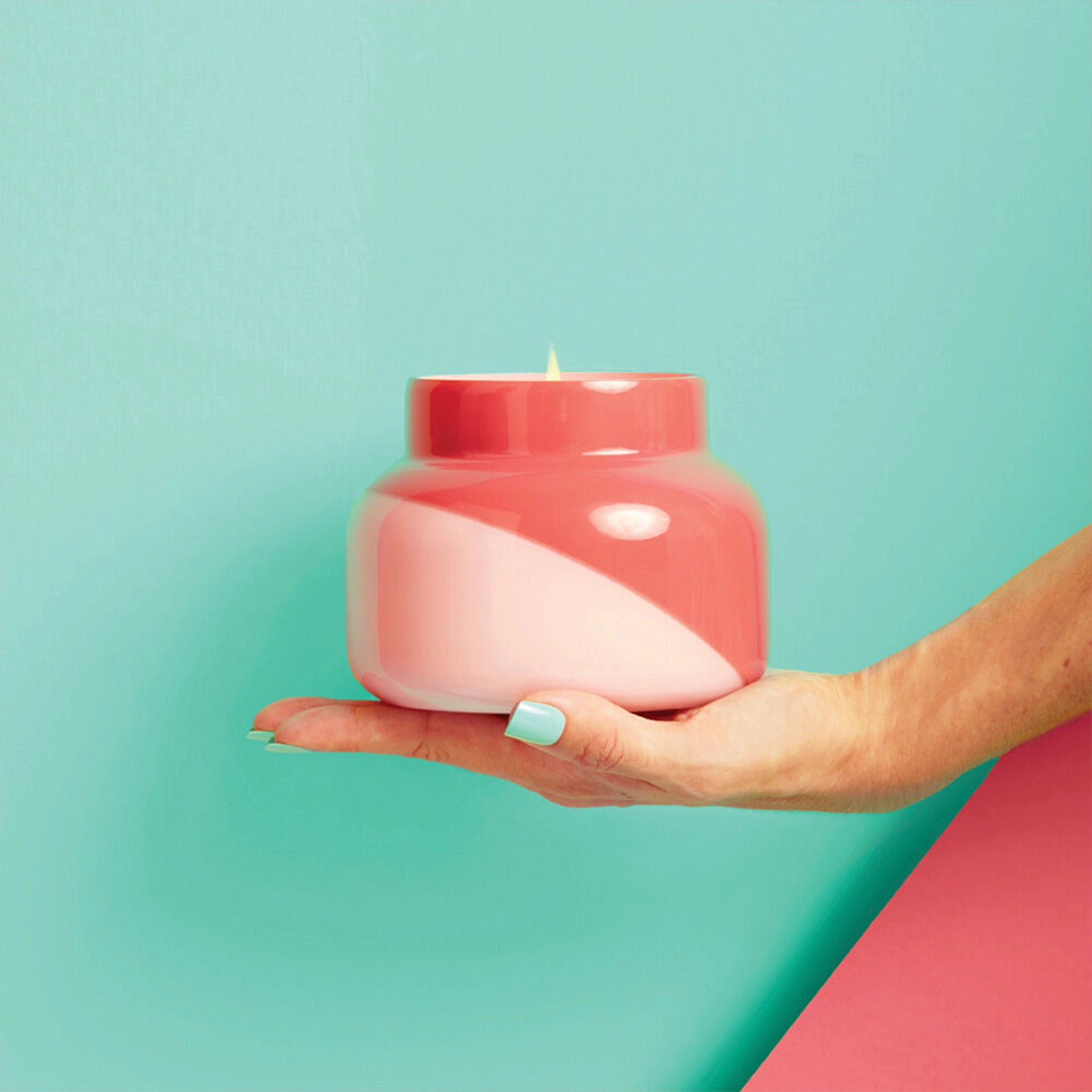 A glass candle jar with a two toned design featuring a light pink and a darker pink that is split diagonally and held in a models hand in front of a teal background.