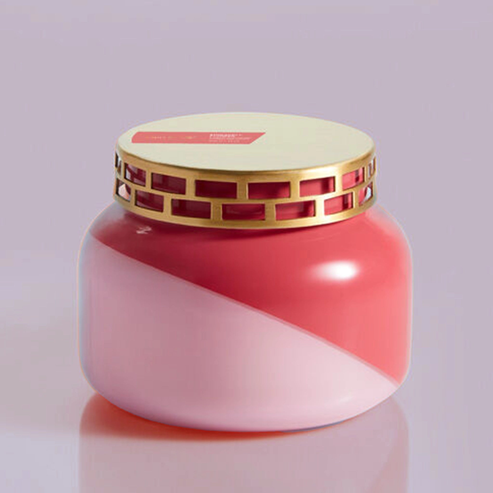 A glass candle jar with a two toned design featuring a light pink and a darker pink that is split diagonally with a gold metal lid with rectangle cut out detailing around the edge.