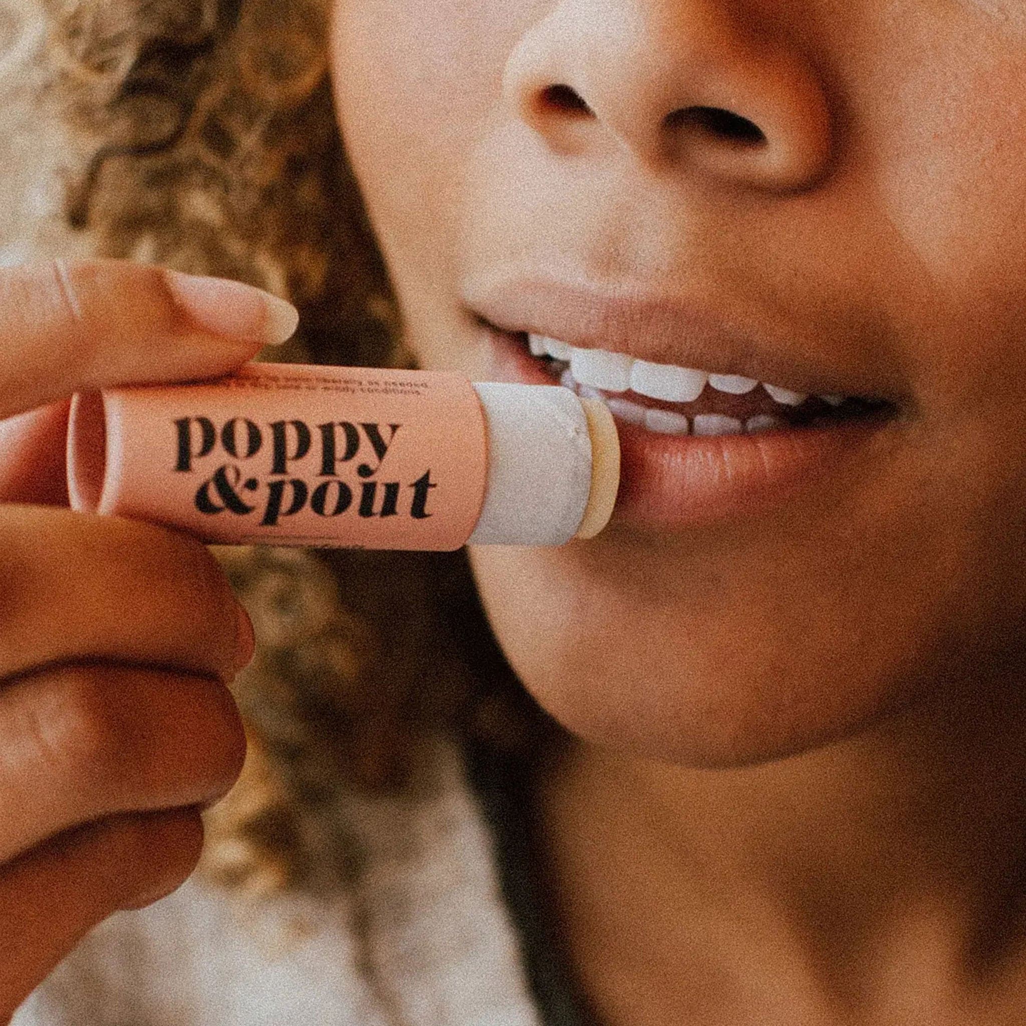 This is a close up picture of a woman’s mouth. She is holding a round peach tube up to her lips. On the left side of the tube is black text that reads ‘poppy &amp; pout.’ 