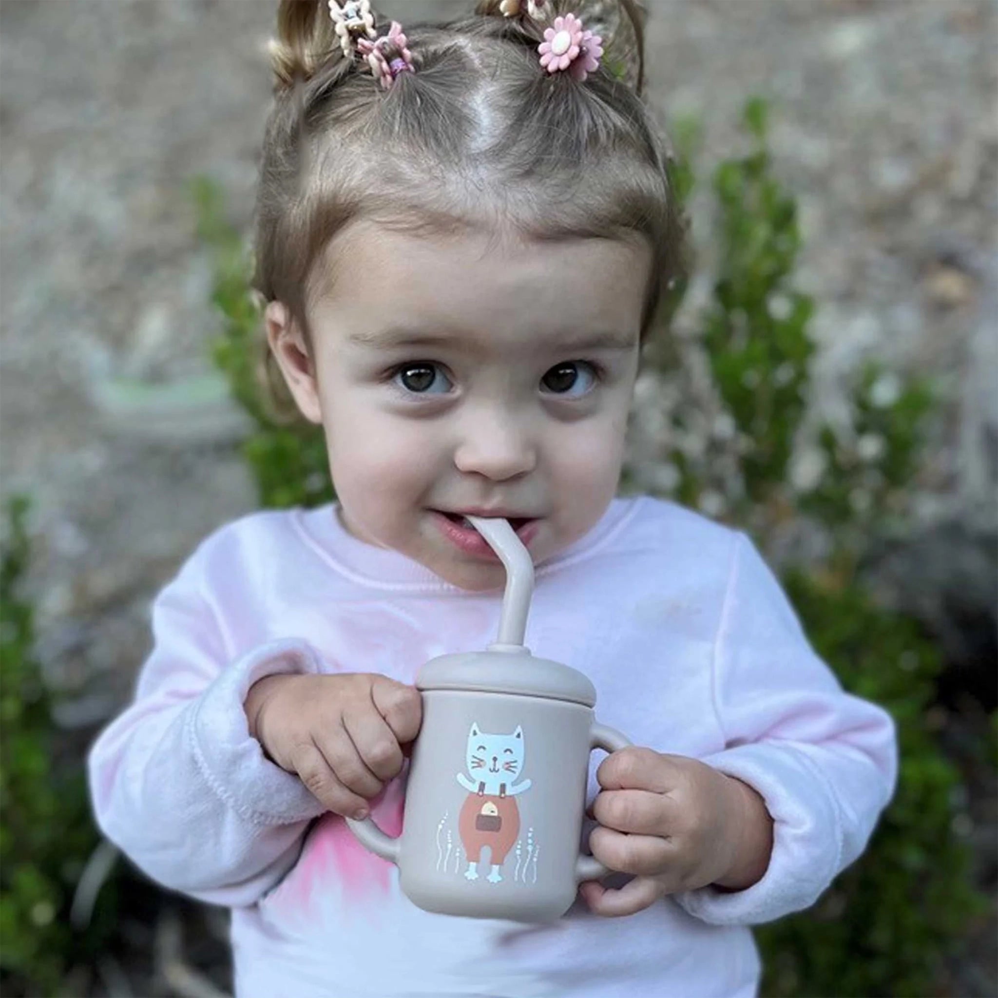 On a cream background is a light pink sippy cup with two handles and straw, designed with a white kitten illustration on the front held by a toddler. 