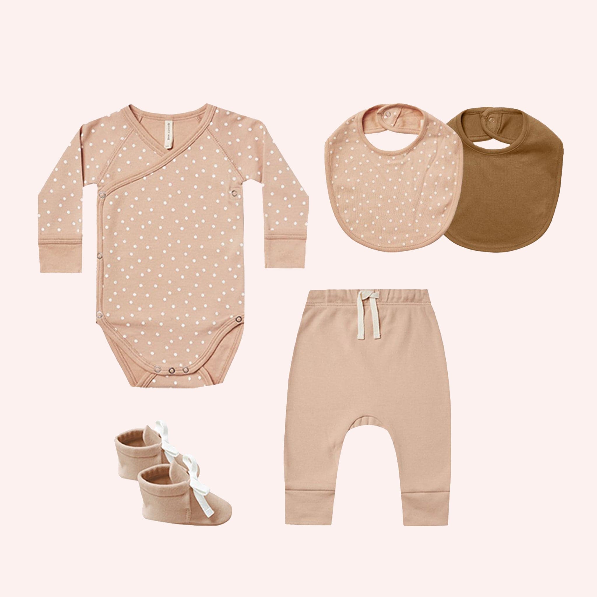 A baby clothing set. A long sleeve onesie in blush pink with white polka dots. A blush bib with white polka dots, a brown bib. Blush pink joggers with white drawstring. Blush booties with white lace ties.