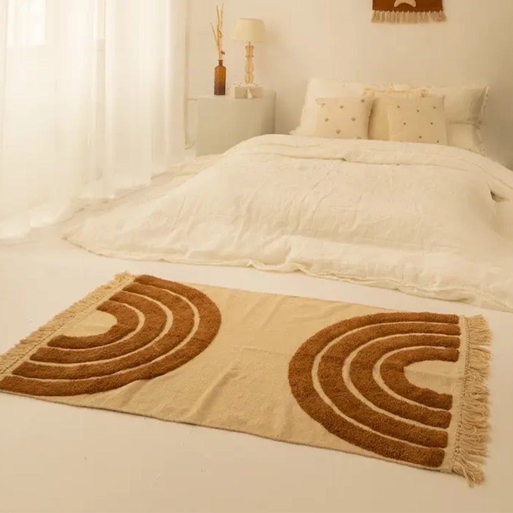 A light beige rug with raised terracotta rainbows on each end of the rug along with a tassel detail on each end as well. 