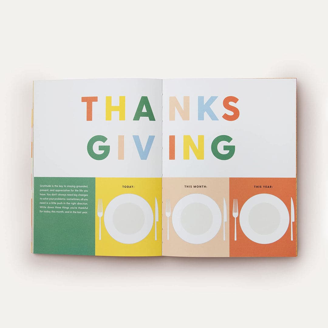 Journal is open and turned to two pages that read &#39;THANKSGIVING&#39; across both pages in orange, yellow, green, peach and baby blue lettering. Below there are four square sections that are labeled &#39;Today&#39;, &#39;This Month&#39;, &#39;This Year&#39; with space to write below in a shape of a Thanksgiving dinner plate.