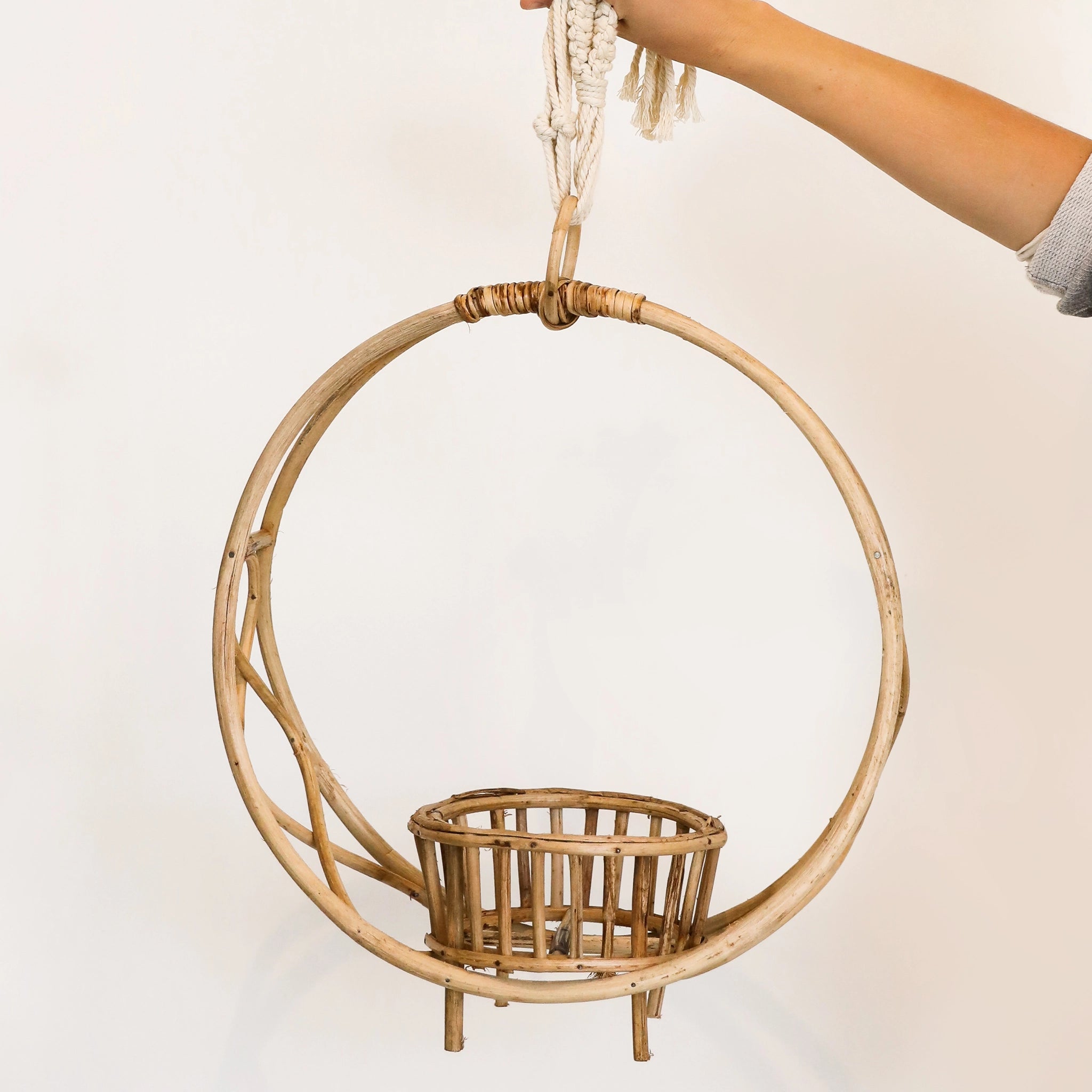In front of a white background is a circle rattan plant holder. On the bottom inside of the holder is a rattan pot to hold the plant. Below the rattan pot is four rattan pegs. At the top of the rattan circle is a small rattan hook.