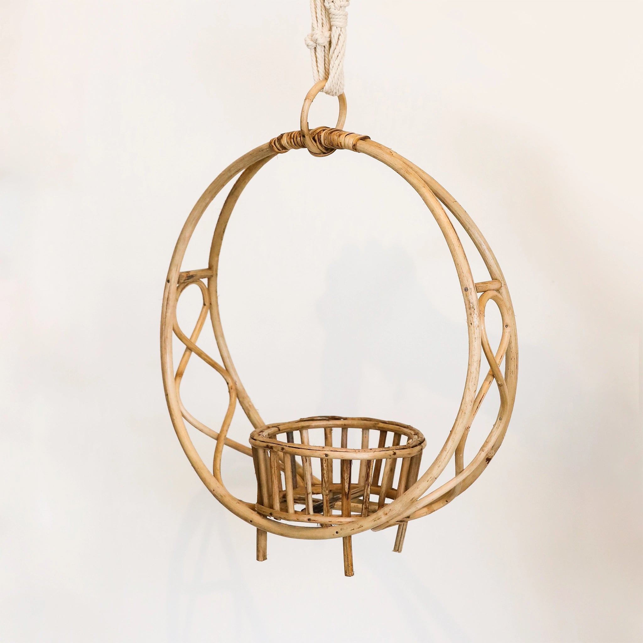 In front of a white background is a circle rattan plant holder. On the bottom inside of the holder is a rattan pot to hold the plant. Below the rattan pot is four rattan pegs. At the top of the rattan circle is a small rattan hook. There is a hand holding a piece of rope that is attached to the hook.