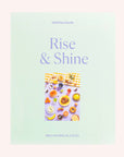 A mint green cardboard boxed puzzle with the words "Rise & Shine" in a light purple text along with an image of an array of bedazzled breakfast items including bagels, fruits, sunny side up eggs and more.
