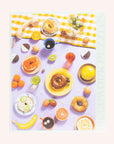 A look at what the completed puzzle looks like which is an array of bedazzled breakfast items including bagels, fruits, sunny side up eggs and more. The primary color tones of this puzzle include shades of light purple, yellow and orange.