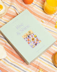 A mint green cardboard boxed puzzle with the words "Rise & Shine" in a light purple text along with an image of an array of bedazzled breakfast items including bagels, fruits, sunny side up eggs and more.