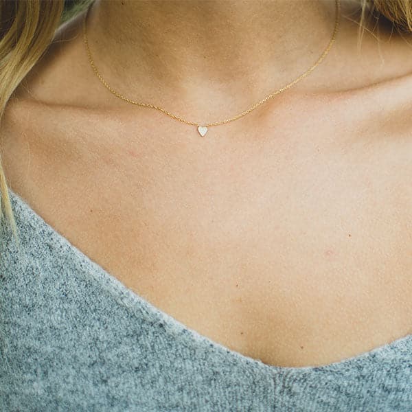 This is a picture of a close up of a model’s neck. The model has blonde hair and is wearing a gray sweater. Around her neck is a thin gold chain with a tiny gold heart in the middle.