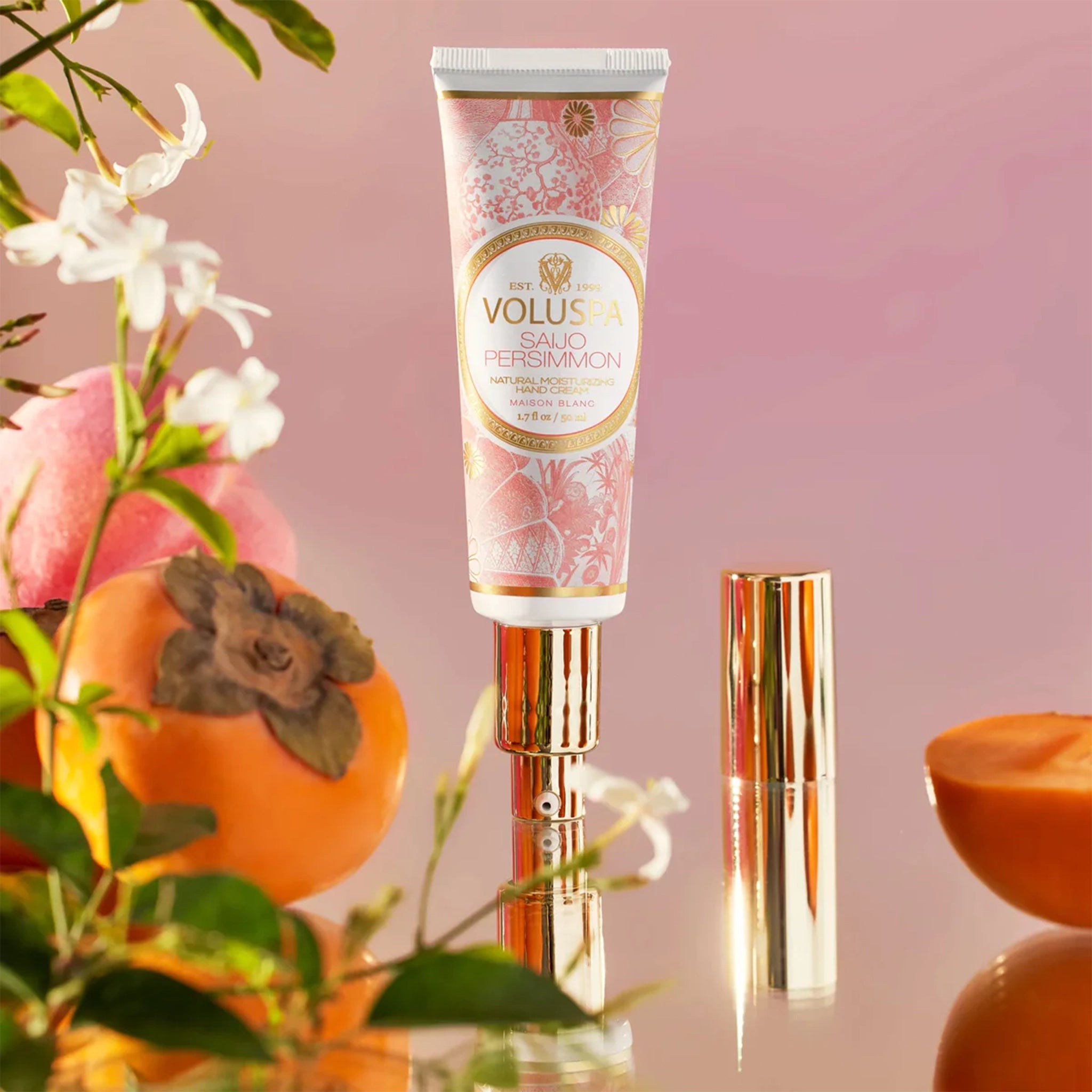 A tube of hand cream decorated with a pink floral pattern with gold text in the center that reads, "Voluspa Saijo Persimmon". The tube of hand cream has a gold pump and gold cap. It is photographed around florals and persimmon fruits.