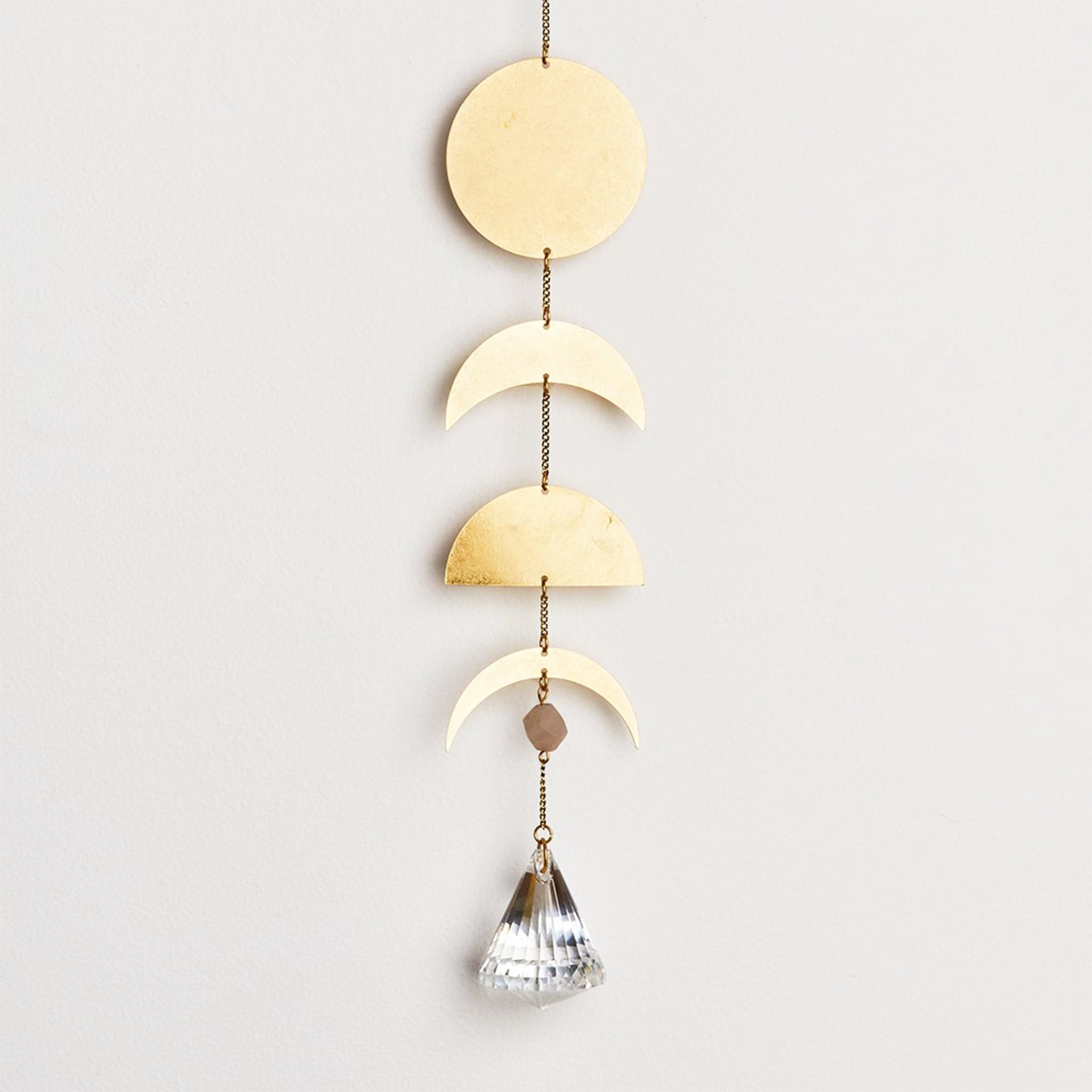 Hanging in front of a white background is a gold chain with a clear, crystal prism at the bottom. Above that is a brown bead. Next is a gold, metal crescent moon shape. Next is a gold, metal half circle. Above that is another gold, metal crescent moon shape just a little thicker. At the top is a gold, metal circle. 