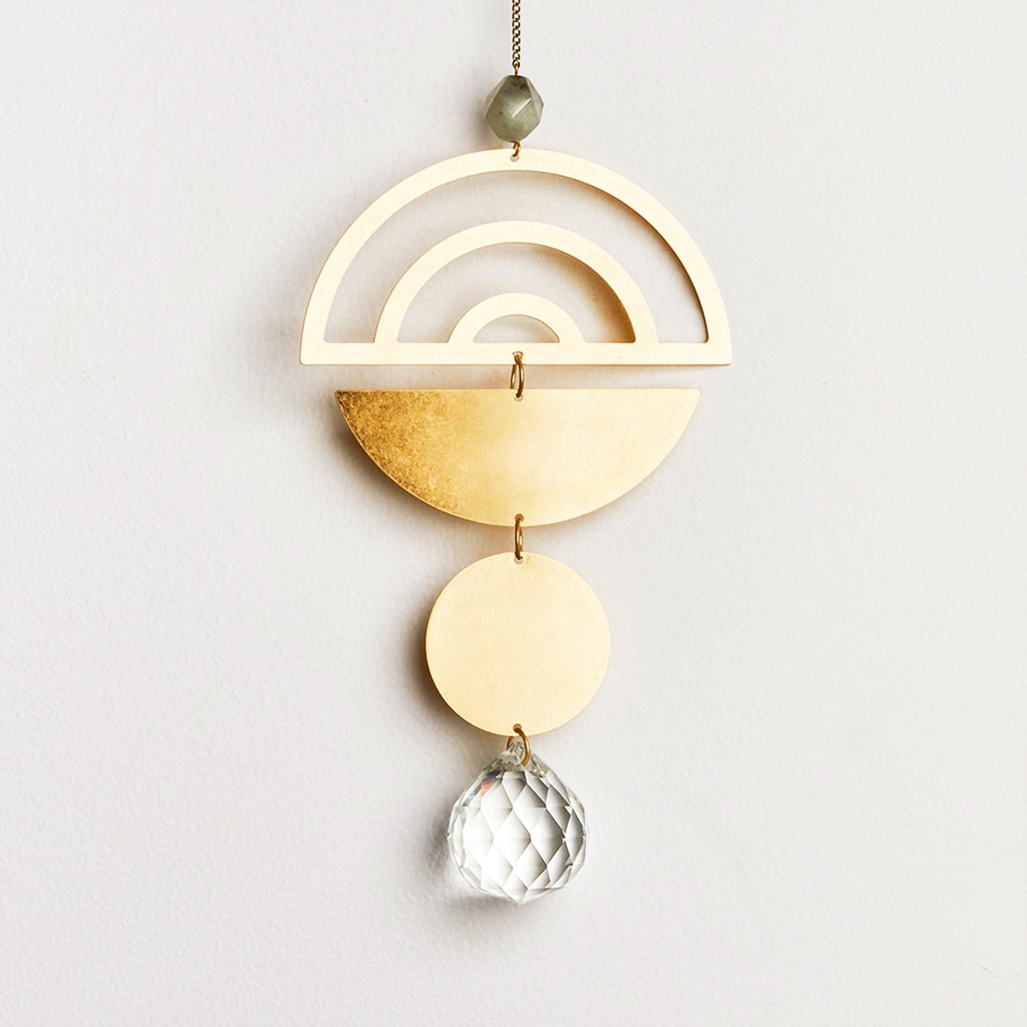 Hanging in front of a white wall is a light catcher. Starting at the top is a dark bead. Next is a gold, metal half circle with the arch at the top. There are three arch cut outs in the half circle. Attached by a small, gold hoop is an upside down, gold half circle. Attached by another gold hoop is a small gold circle. At the bottom is a circle, clear crystal. 