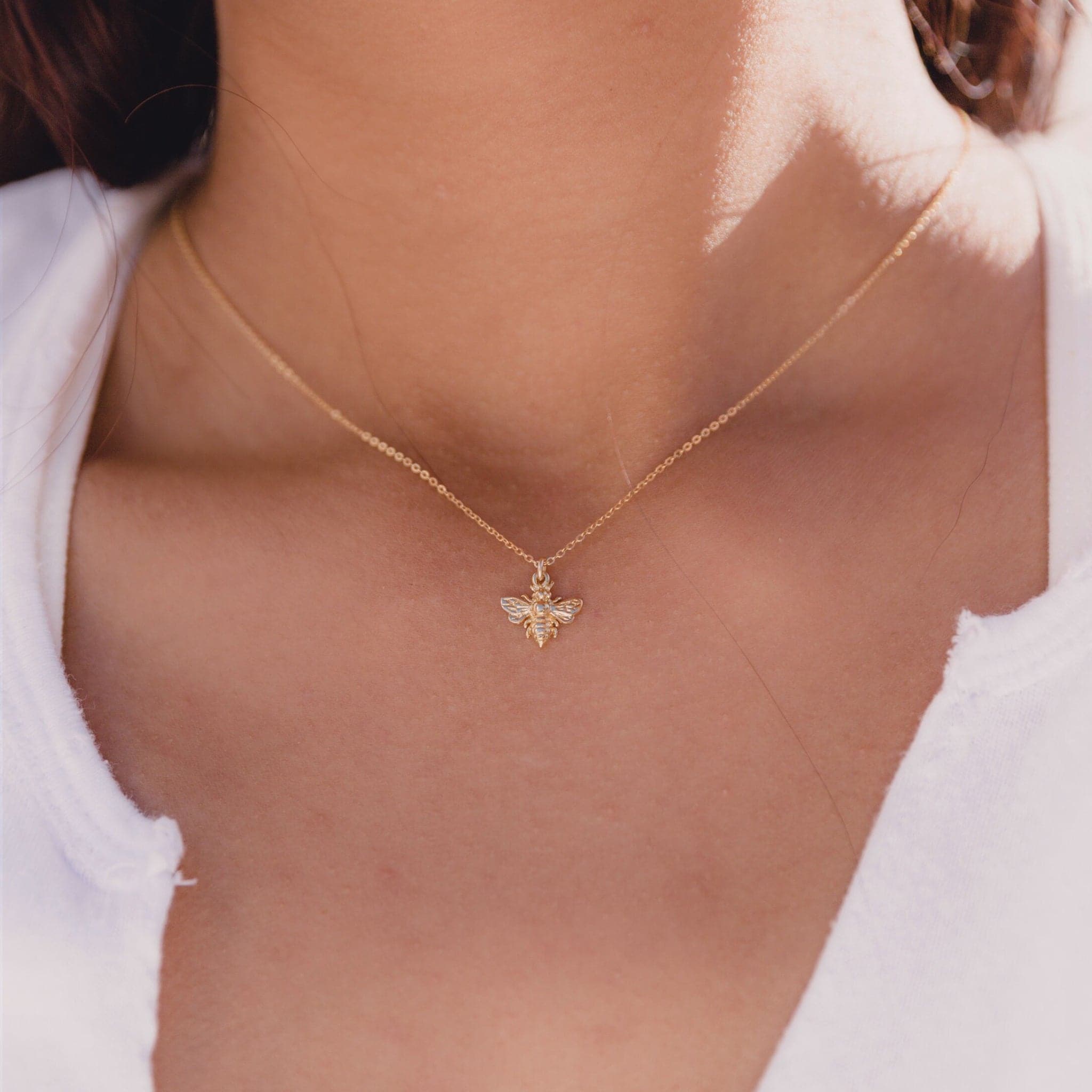 This is a picture of a woman’s neck. She is wearing a white shirt. Around her neck is a thin gold chain with a small gold bee charm in the middle. 