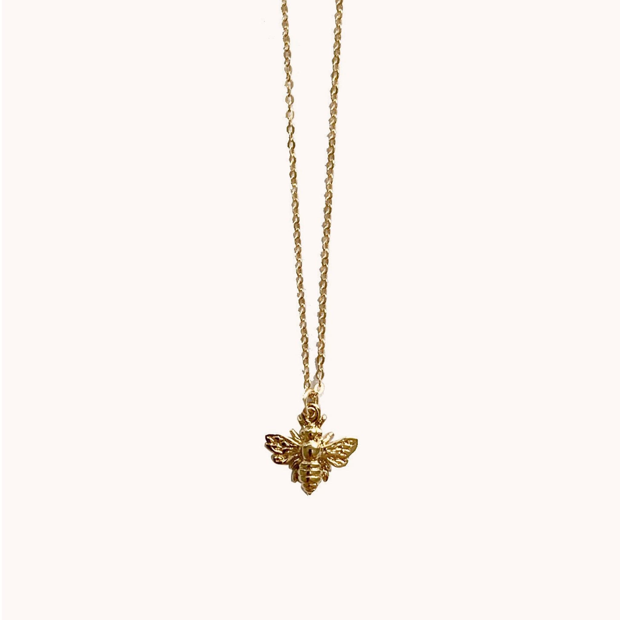 Hanging in front of a white background is a gold chain necklace with a small gold bee charm in the middle. 