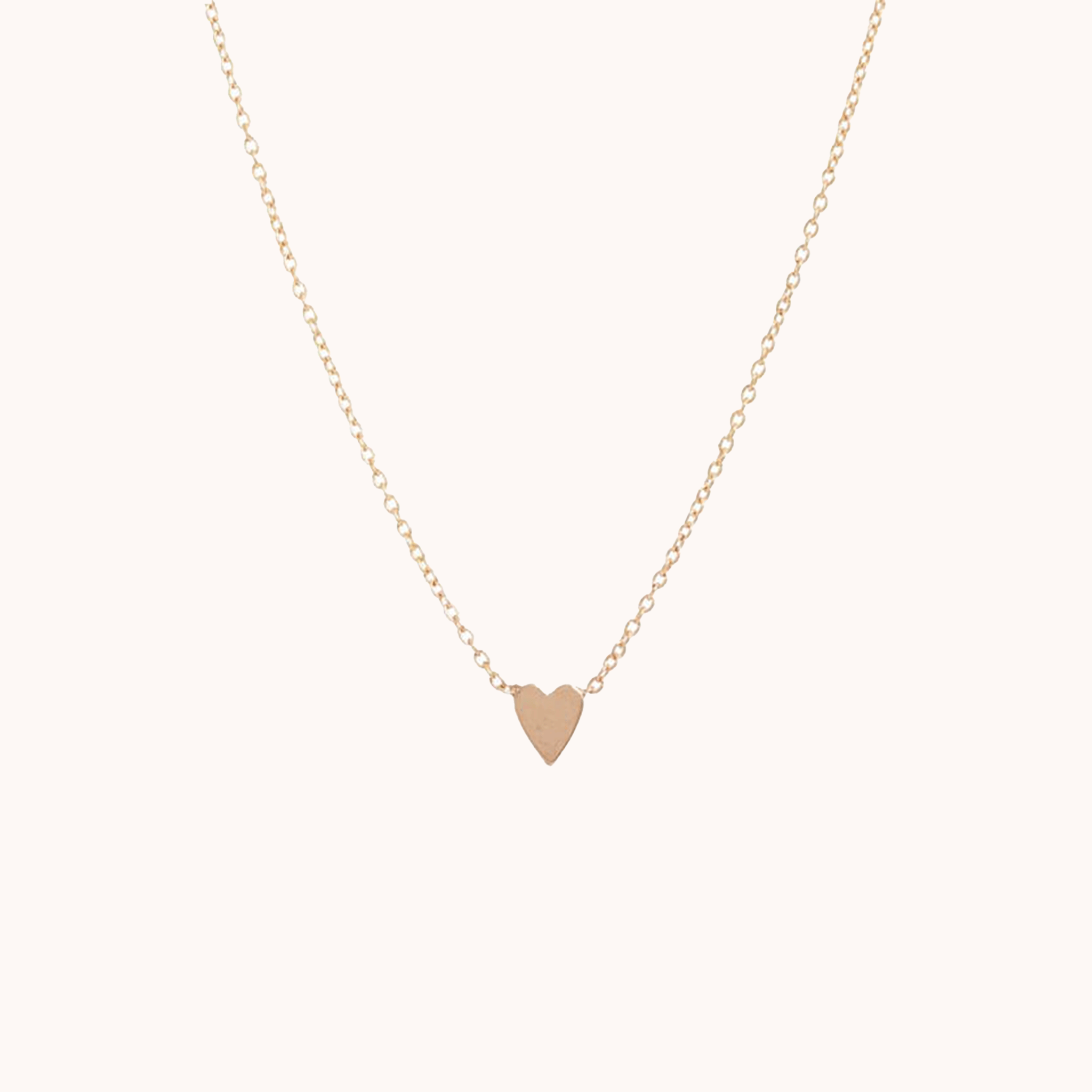 In front of a soft pink background is a thin gold chain with a thin gold heart in the middle.