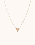 In front of a soft pink background is a thin gold chain with a thin gold heart in the middle.