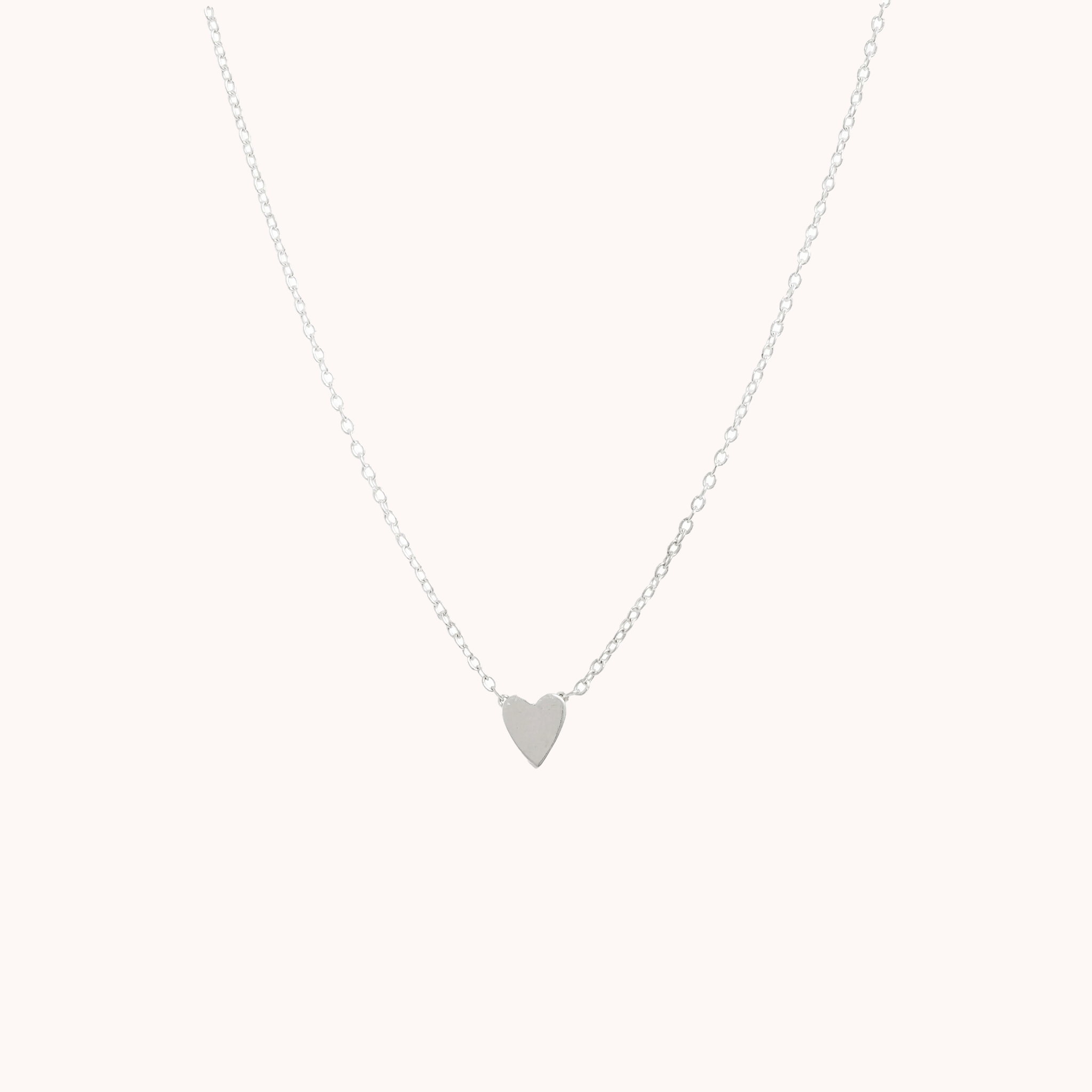 In front of a soft pink background is a thin silver chain with a thin silver heart in the middle.
