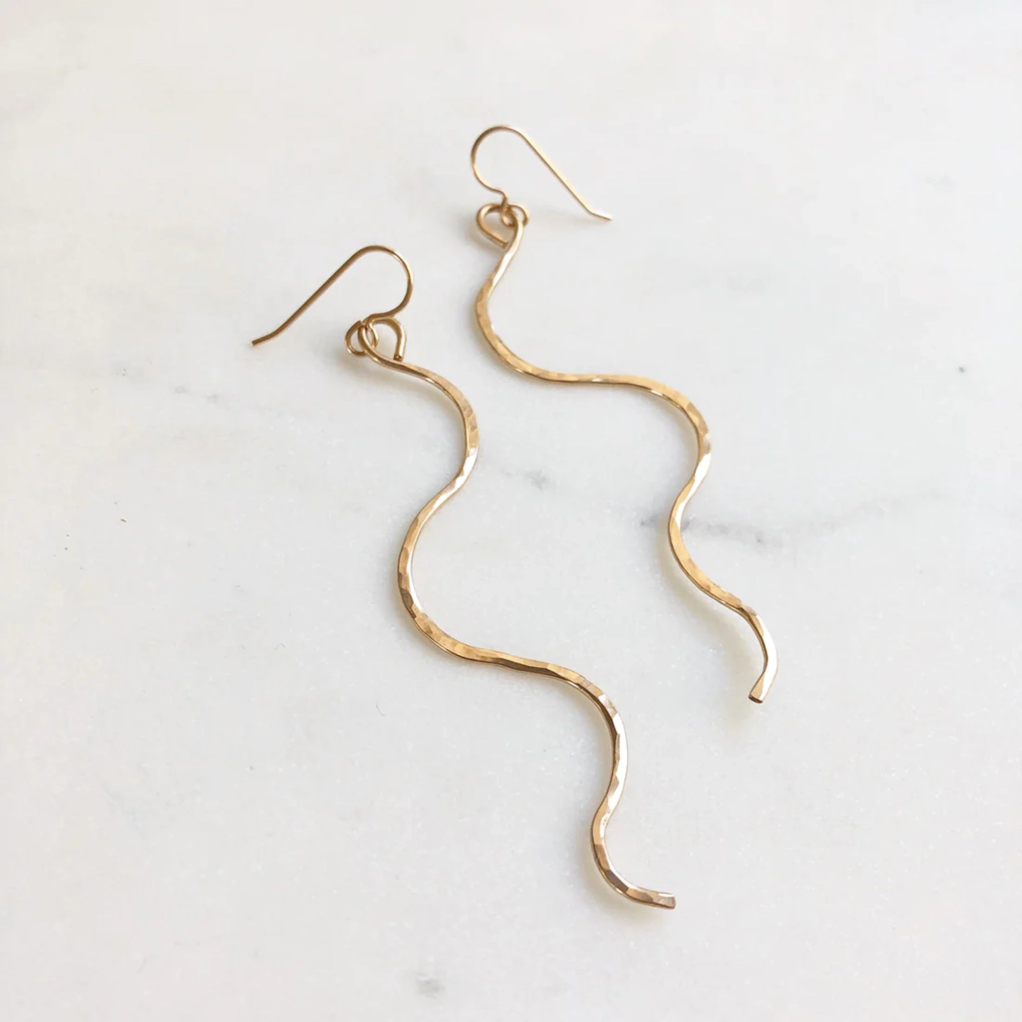 Two thin gold dangle earrings in a wavy "serpent like" shape with a hook earring back and a lightly hammered texture. 