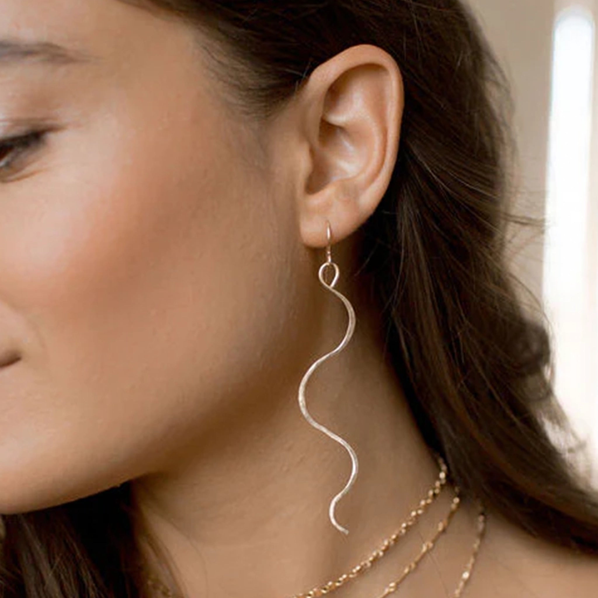 Two thin gold dangle earrings in a wavy "serpent like" shape with a hook earring back and a lightly hammered texture worn on a model and come down to about her mid neck.