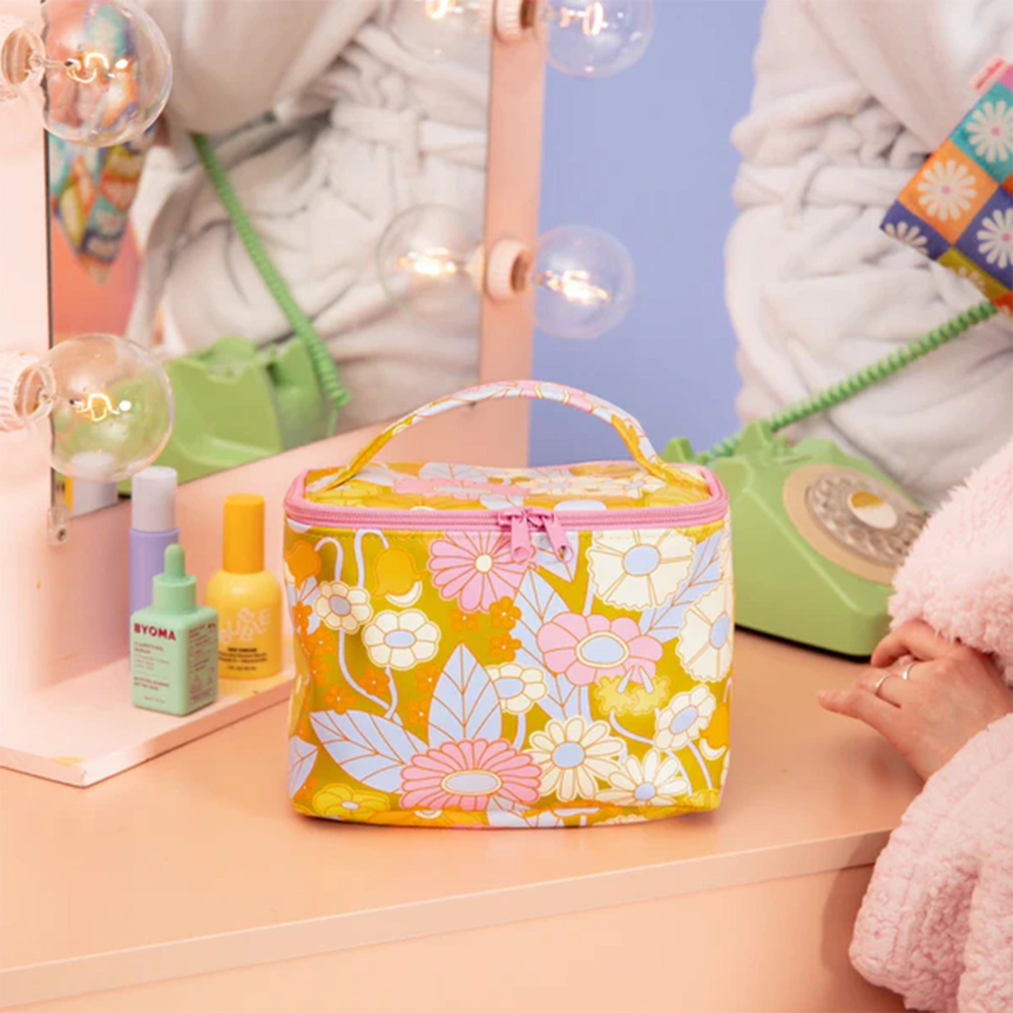 A colorful floral printed cosmetics bag with a flat top and bottom, curved edge and a single strap on top as well as a zip around closure. The floral design features shades of olive green, creams, pink, and light blue.