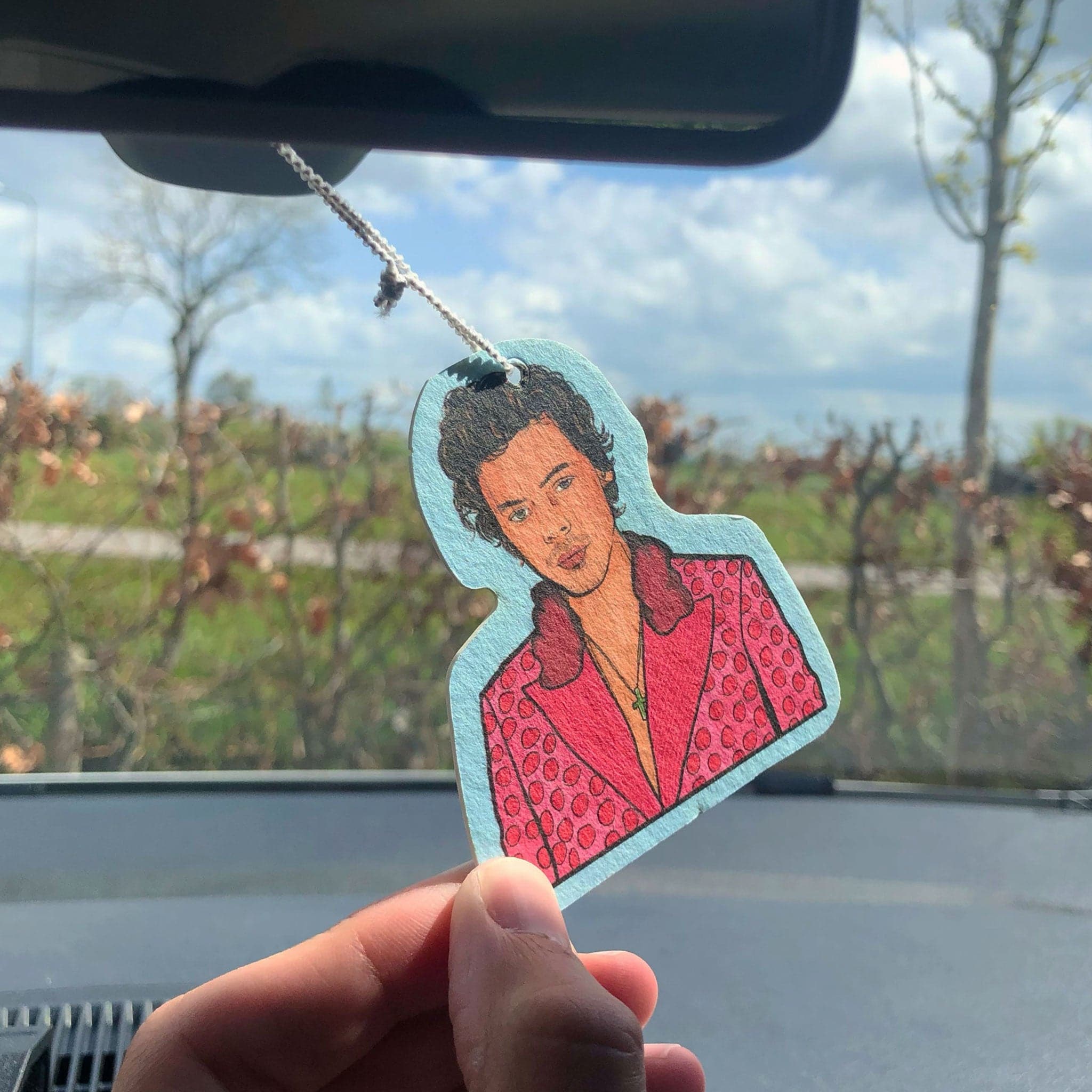 Light aqua air freshener with display the top half of Harry Styles wearing an iconic pink polo dot suit and cross necklace. Air freshener hangs on a white elastic loop from car mirror. A hand holds the air freshener in the light of the car window. 