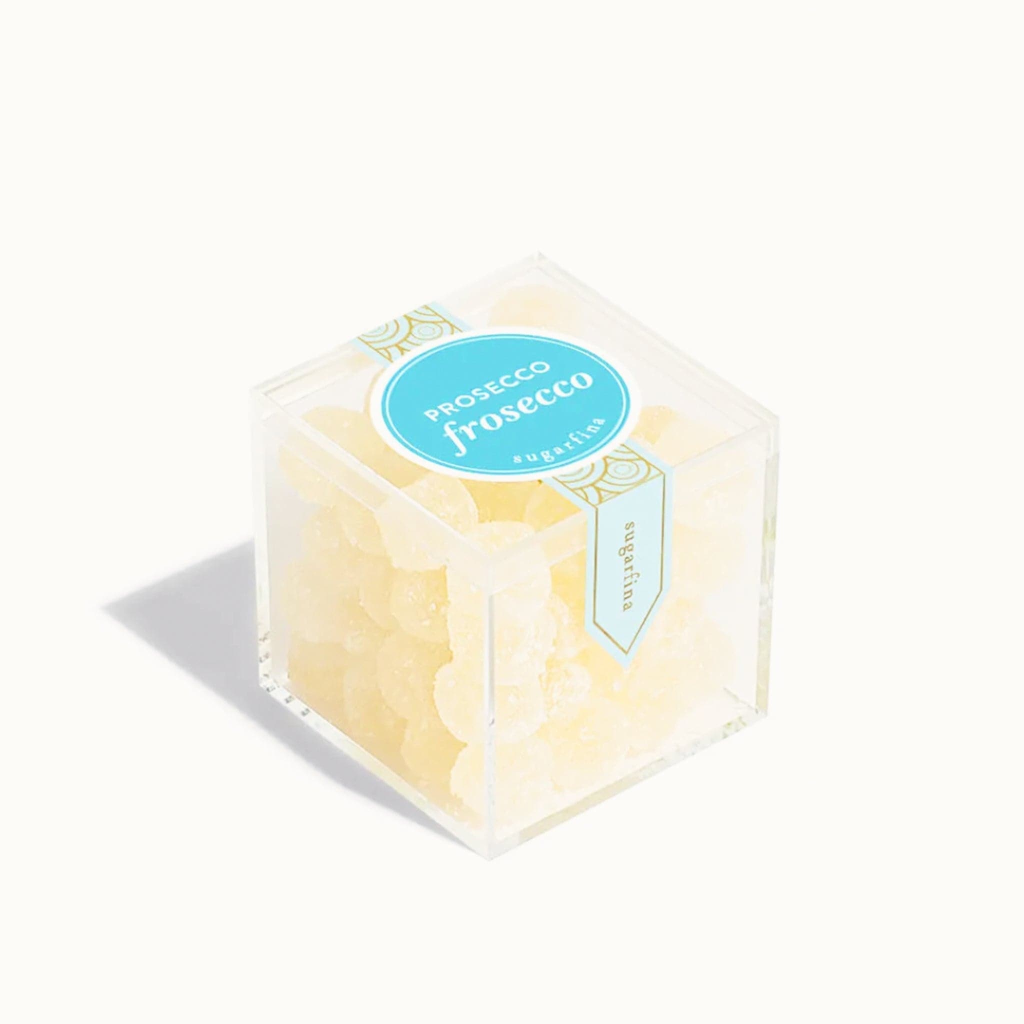 In front of white background is a clear cube container. It is filled with light yellow candies that are coated in with sugar. On top is a bright blue circle sticker with white text in the middle that reads ‘prosecco frosecco.’ 