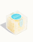 In front of white background is a clear cube container. It is filled with light yellow candies that are coated in with sugar. On top is a bright blue circle sticker with white text in the middle that reads ‘prosecco frosecco.’ 
