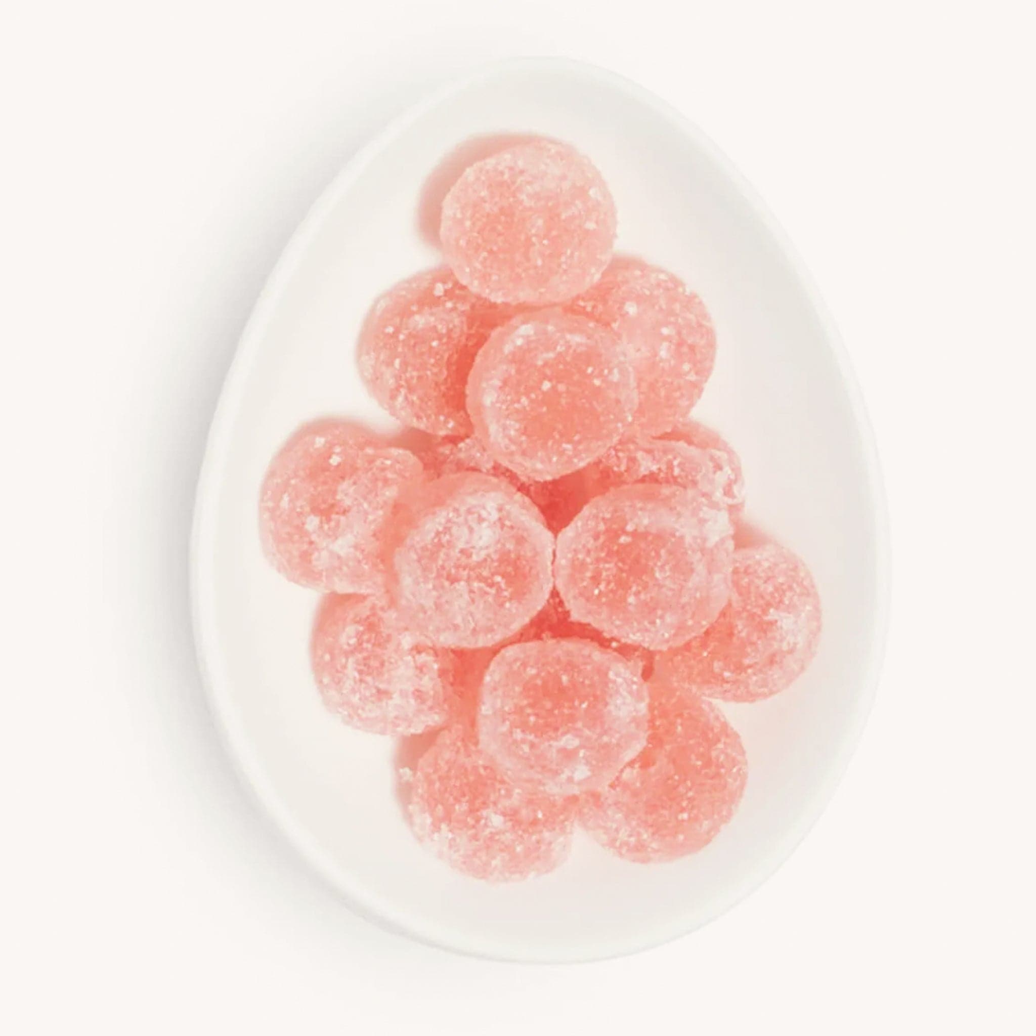 On a white background is a white egg shaped dish with round light pink candies dusted with white sugar. 