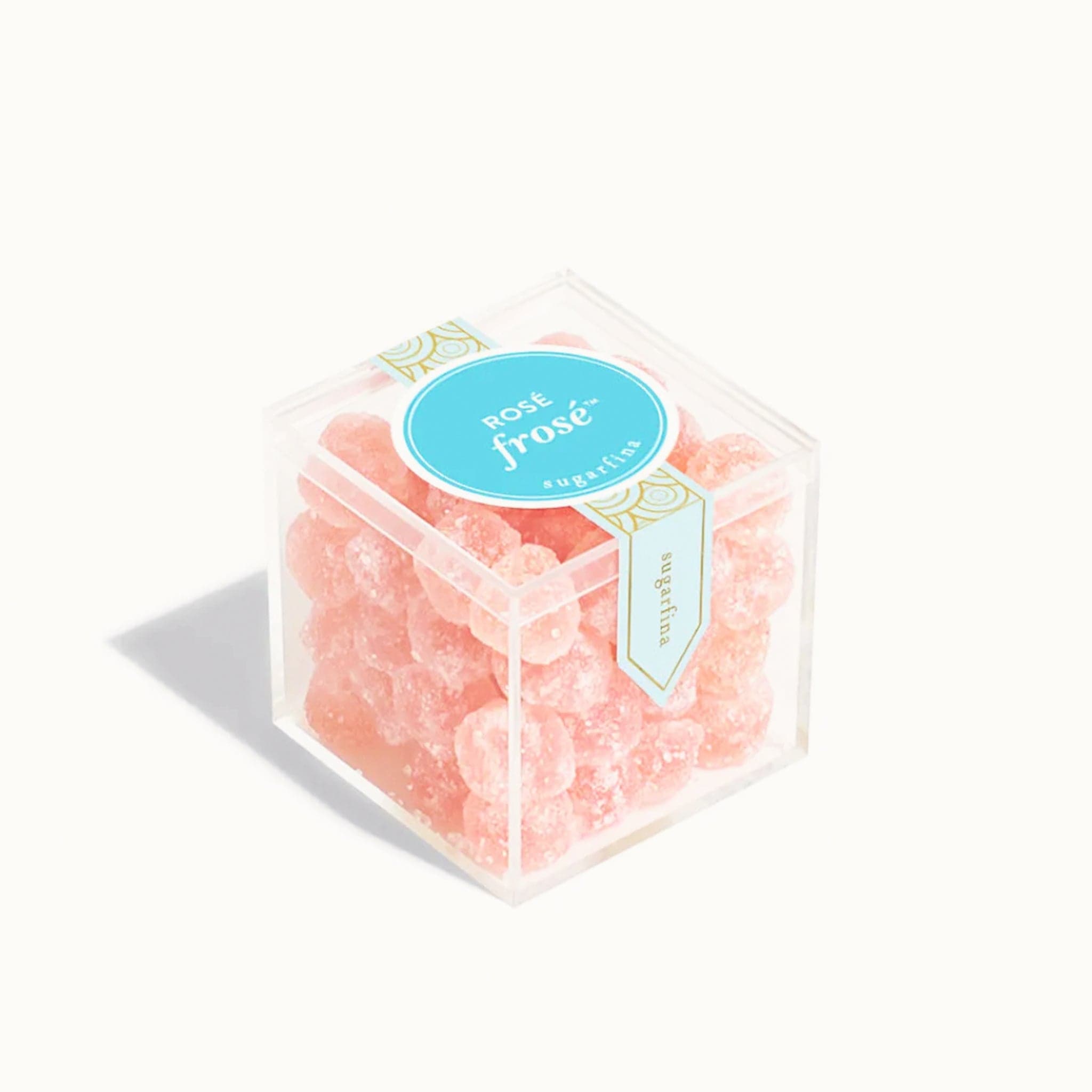 In front of a white background is a clear plastic cube filled with round light pink candies dusted with white sugar. On top is a vertical light blue sticker with gold circles on it. In the middle of the top is a bright blue circle sticker. Inside the circle is white text that reads ‘rosé frosé.’ 