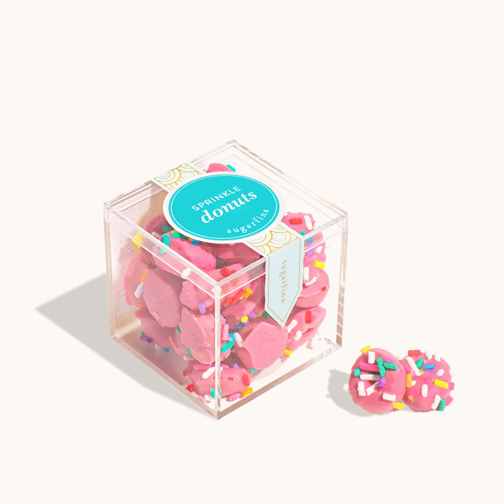 Plastic cube of miniature pink donut candies topped with colorful sprinkles. The box reads &#39;Sprinkle donuts&#39; across a teal circular logo. Two donut candies are placed in front of the box. 