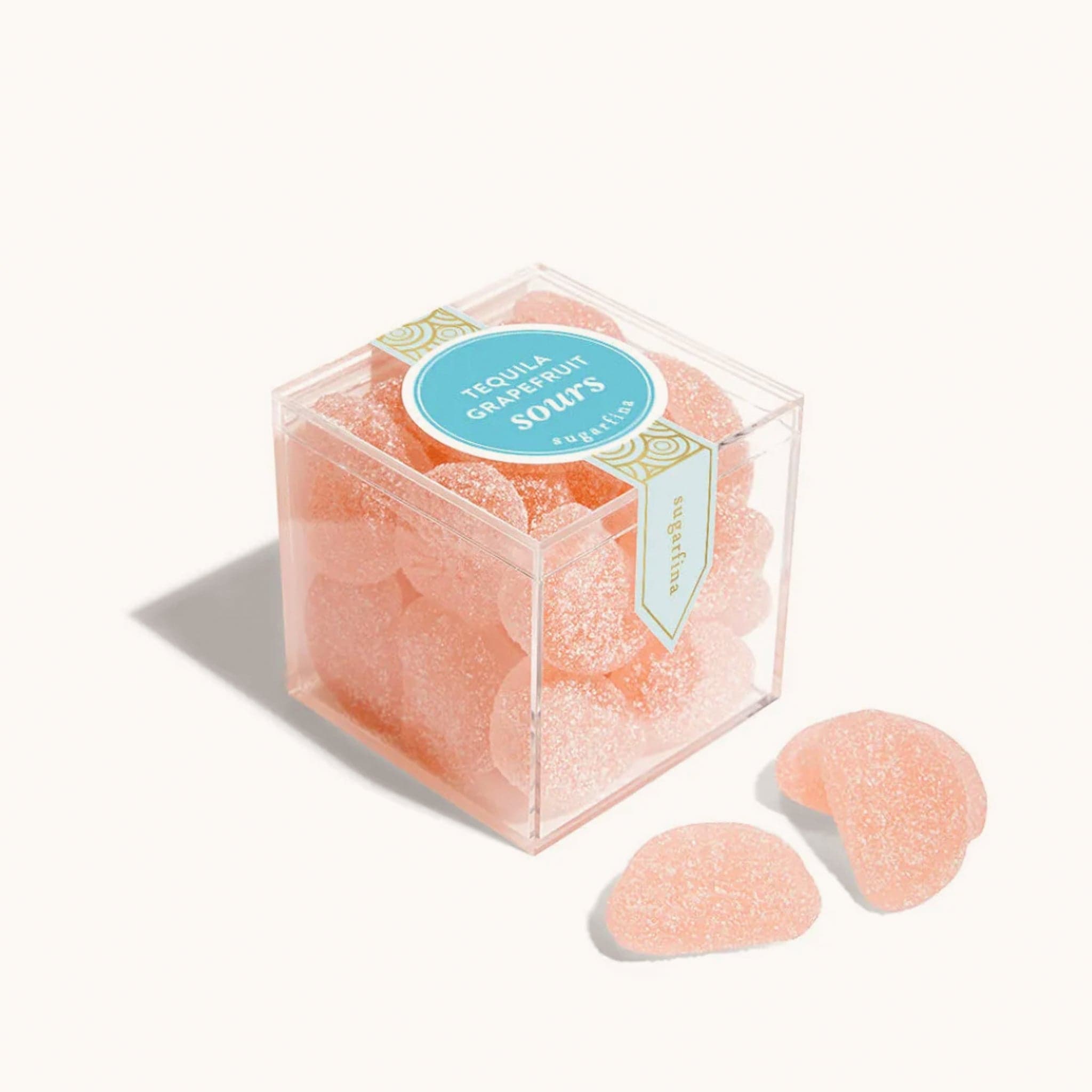 Plastic cube box filled with grapefruit gummies covered in sugar crystals. The clear box reads 'Tequila Grapefruit Sours' on a deep teal label. Three pieces are placed in front of the box.  