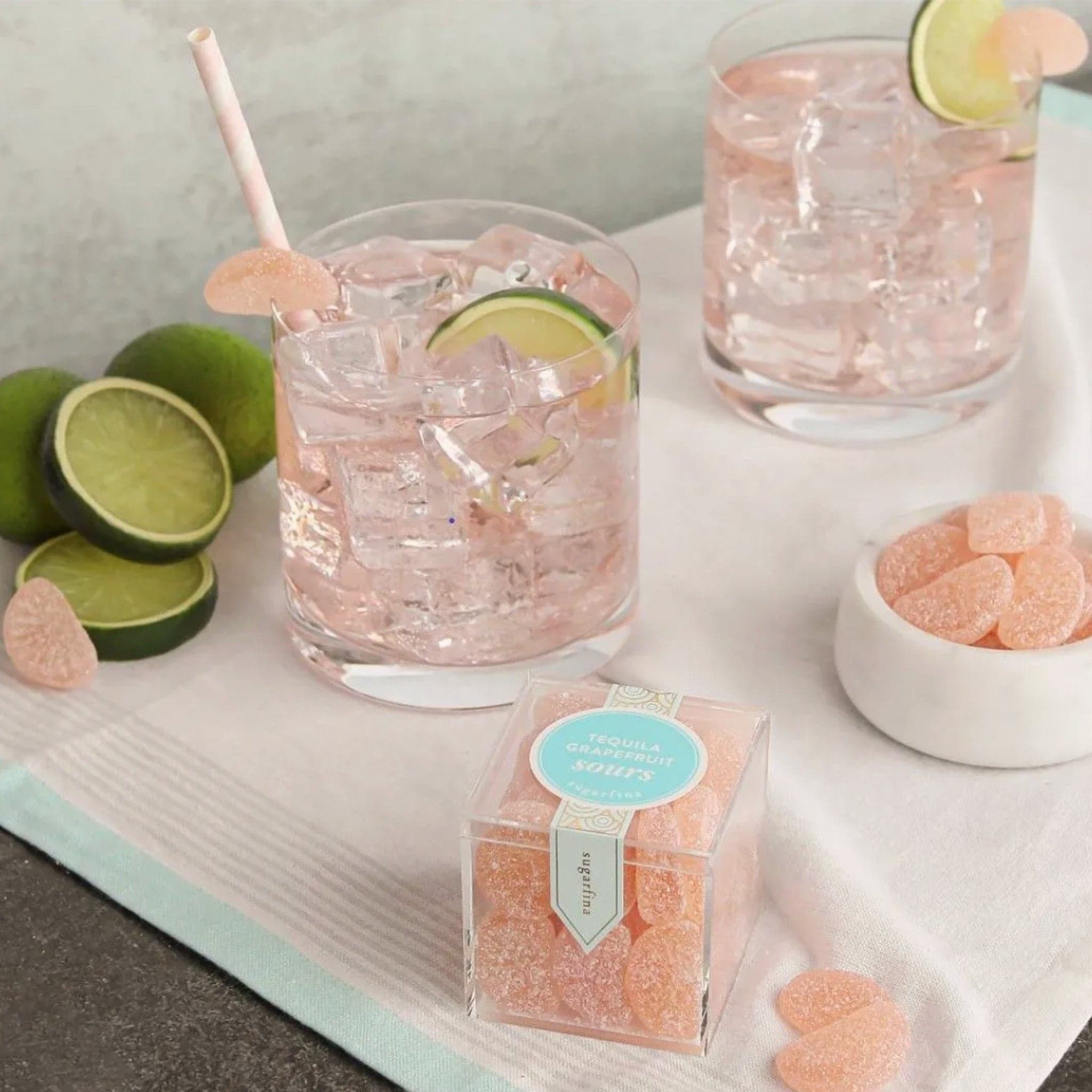 Plastic cube box of grapefruit gummies positioned on a soft pink cocktails garnished with gummies and limes. A small bowl filled with gummies is positioned next to the pair of glasses. 
