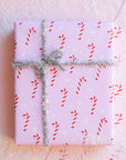 A single sheet of gift wrap with a blush background and a repeating red and white candy cane print. The gift wrap is reversible and the other side features a light purple / lavender background with an all over red and white candy cane design but the candy canes are slightly smaller than the reverse. Present is wrapped with a silver tinsel ribbon and surrounded by snow.