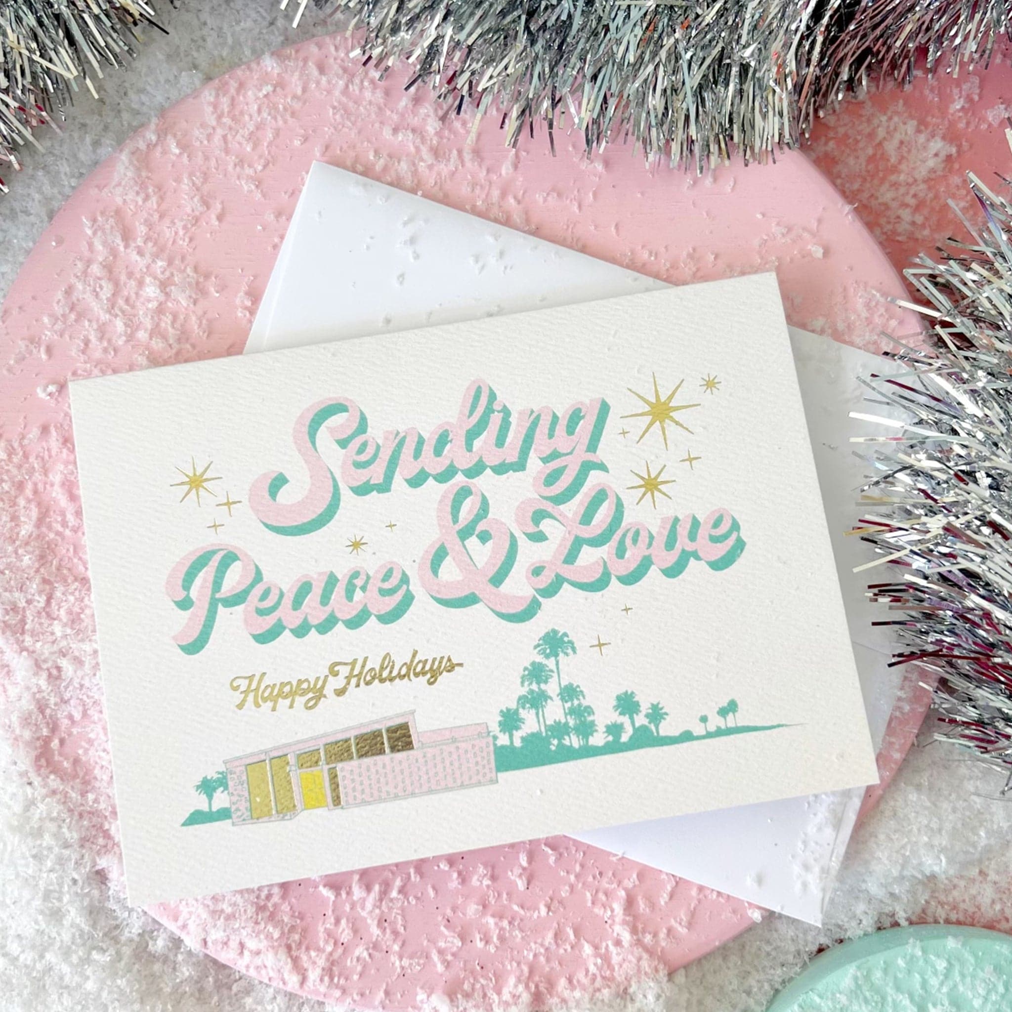A cream card with pink letters outlined in turquoise that read, "Sending Peace & Love" along with a graphic of a midcentury house in front of desert palms and gold foiled letters that read, "Happy Holidays". 